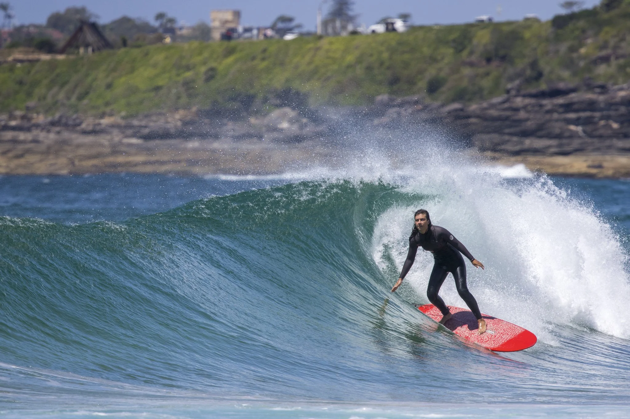 A woman in a black wetsuit surfing on a red surfboard on a wave near the shoreline with a rocky coast and houses in the background.