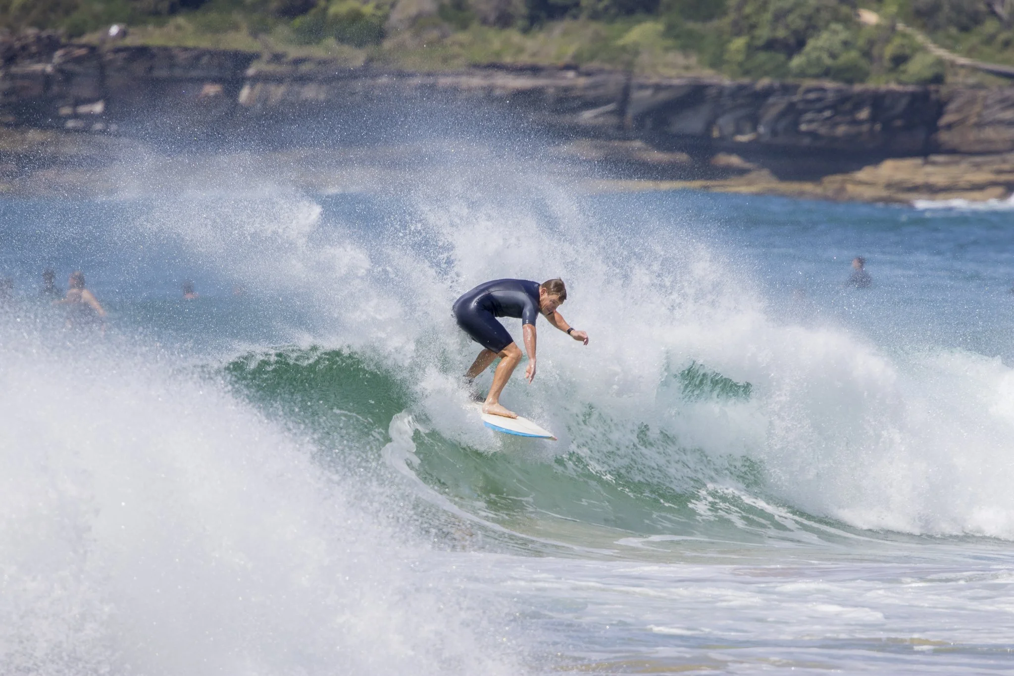 A person surfing on a wave in the ocean with other surfers in the background and a rocky shoreline in the distance.