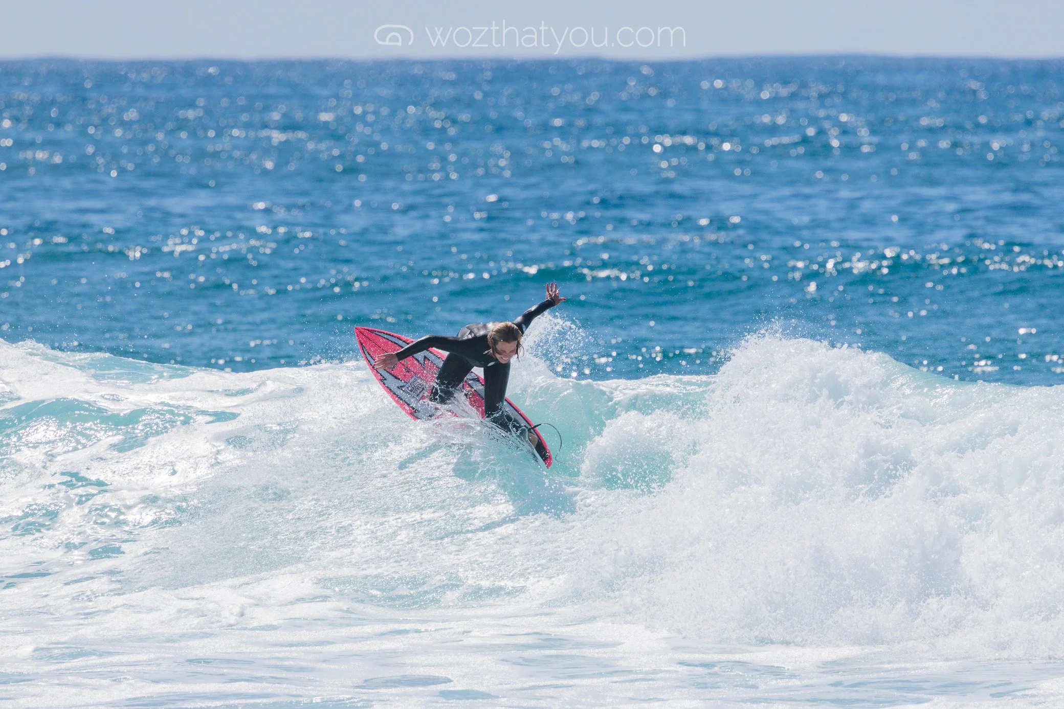A person surfing on a pink surfboard in the ocean, wearing a black wetsuit, riding a wave with a blue ocean background.