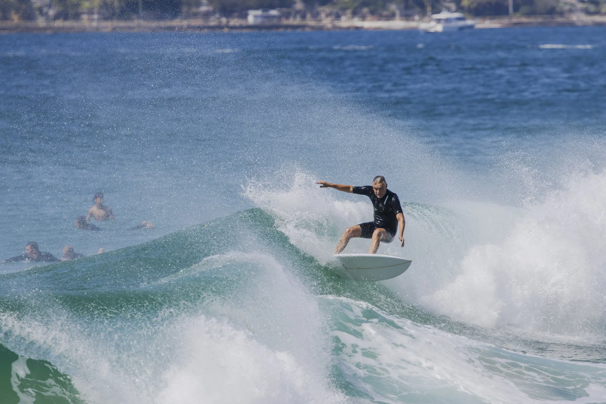 A man is surfing on a wave in the ocean with several people in the background, and a boat is visible on the water.