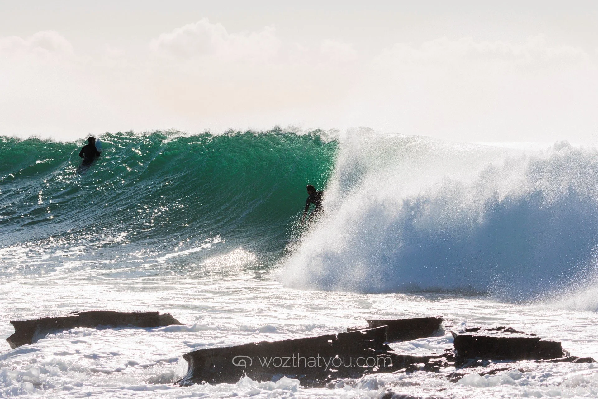 Two surfers riding a large wave on the ocean, with rocky shoreline in foreground and cloudy sky above.