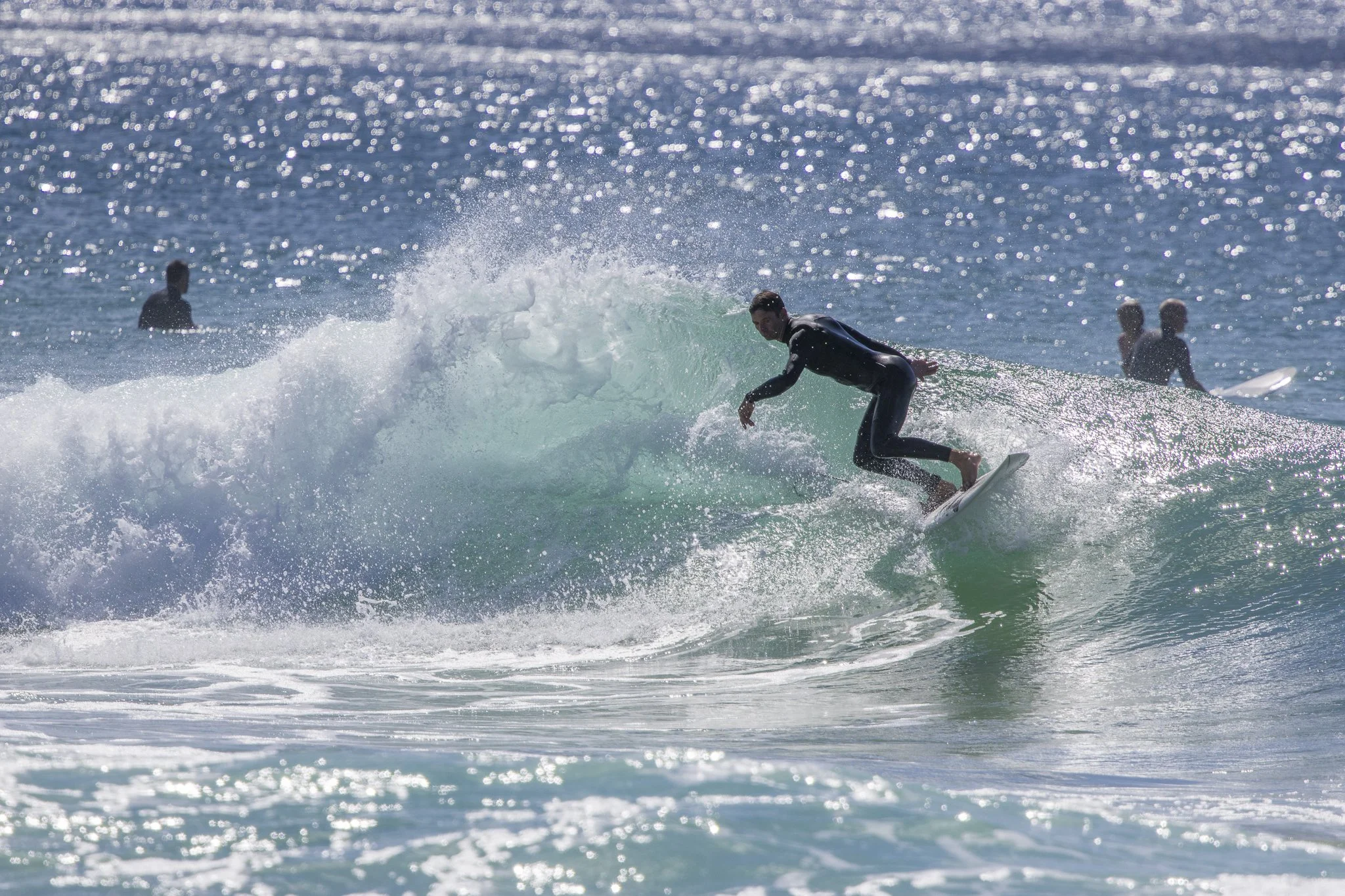 A surfer wearing a black wetsuit riding a green wave with several other surfers in the background in the ocean.