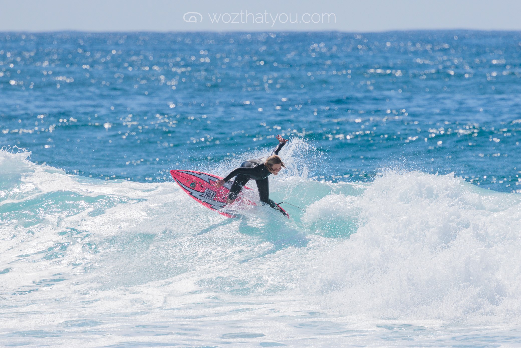 A woman surfing on a pink surfboard in the ocean with blue water and white waves.