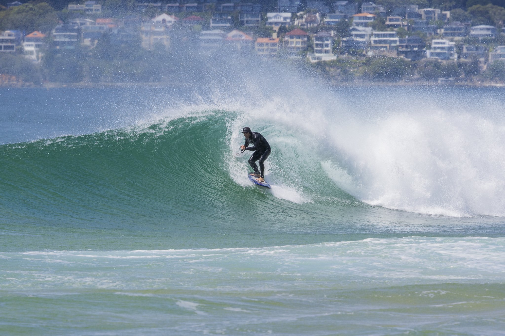 Surfing in a black wetsuit on a green wave near a coastal town with houses in the background.