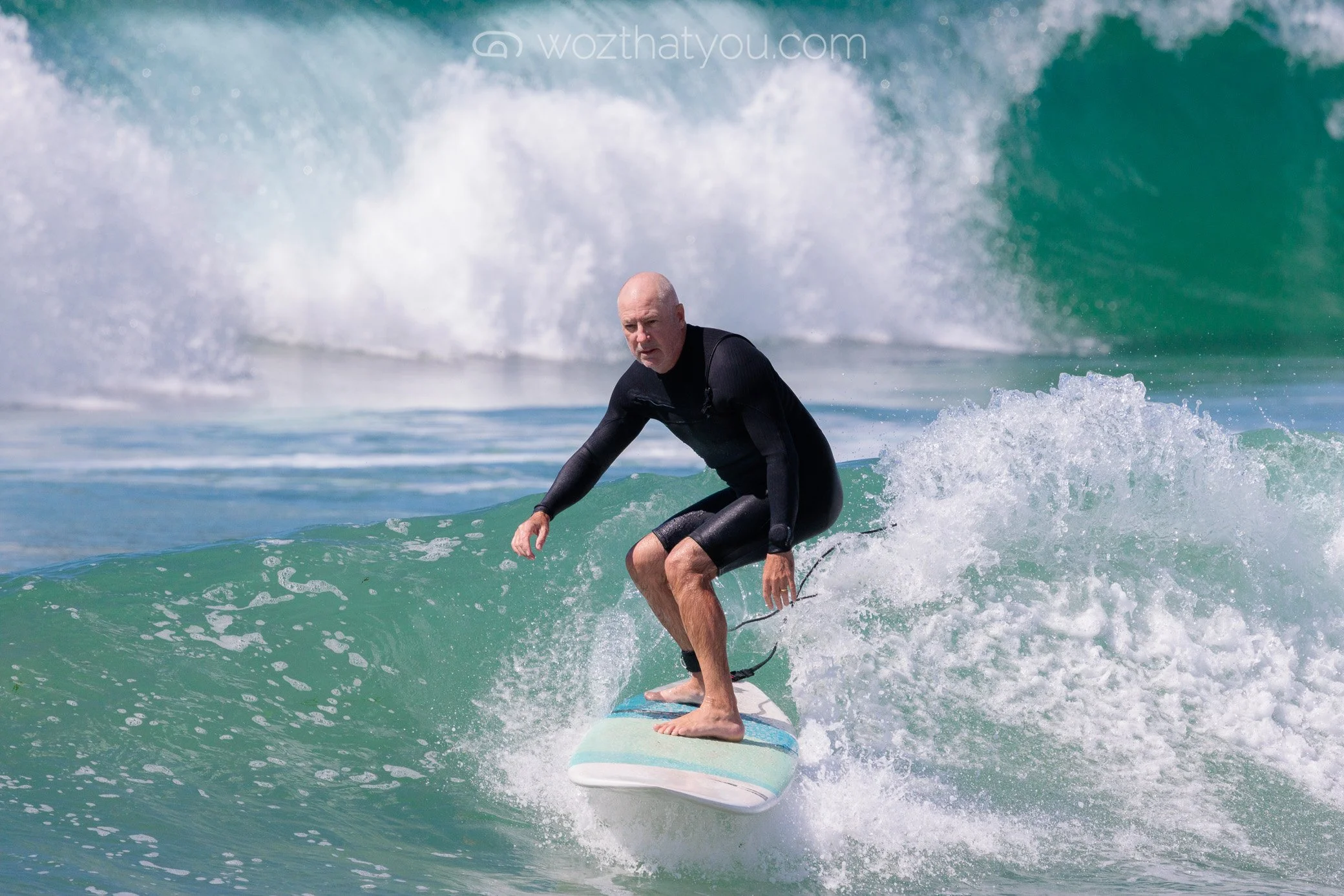 A man with a shaved head surfing on a wave in the ocean, wearing a black wetsuit.