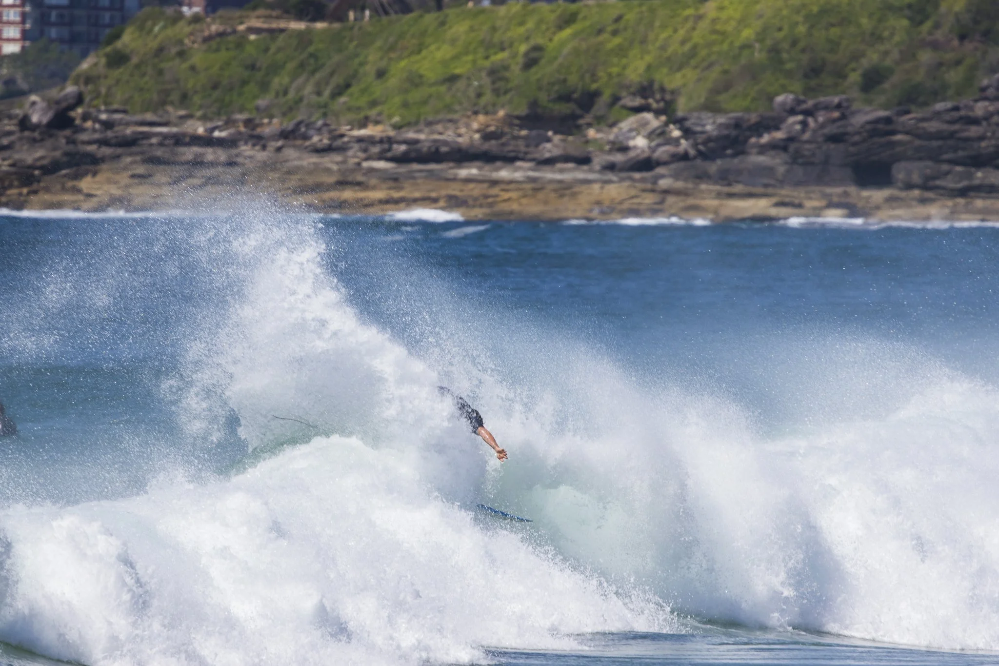 A person falling off a surfboard into the ocean waves near a rocky shoreline with greenery.