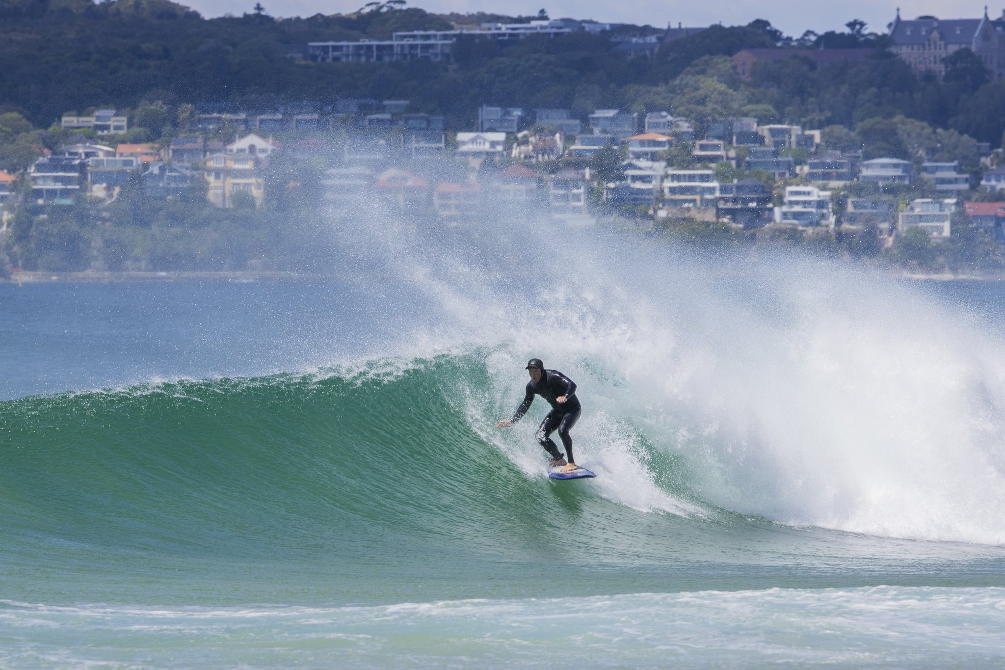 A person surfing on a wave near a coastal town with houses on a hillside in the background.