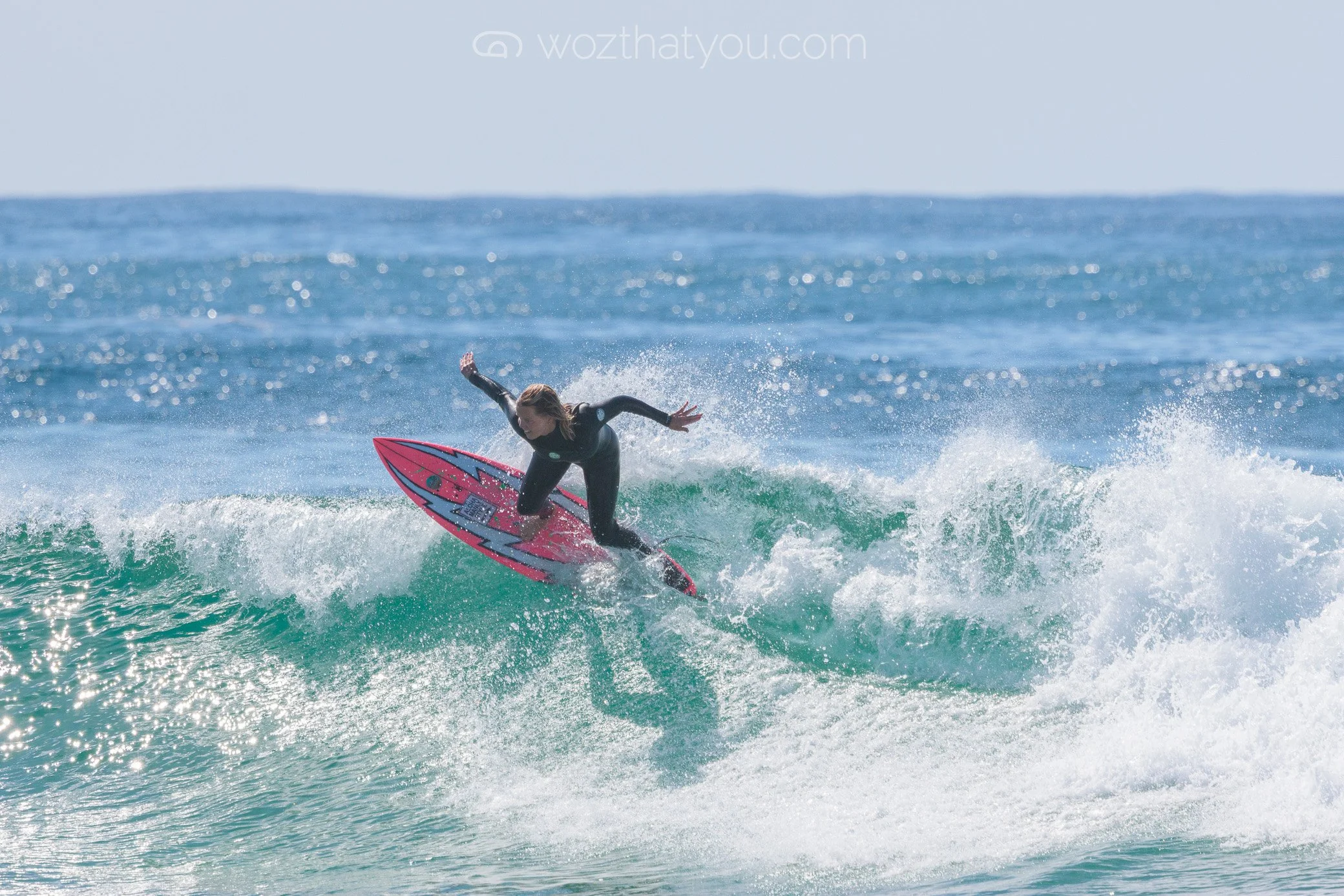 A woman in a black wetsuit surfing on a pink surfboard on a green wave in the ocean.