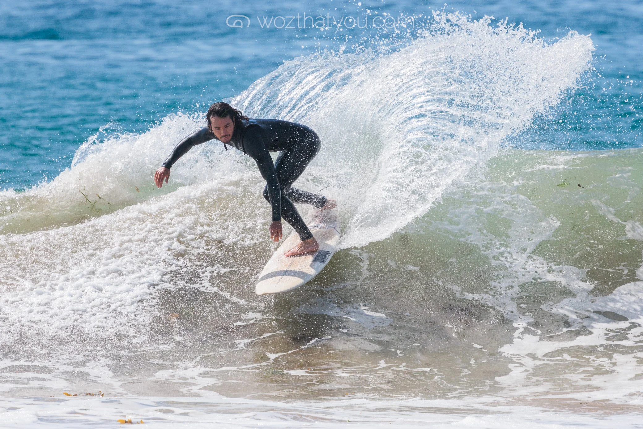A person in a black wetsuit surfing on a white surfboard on a wave at the beach.