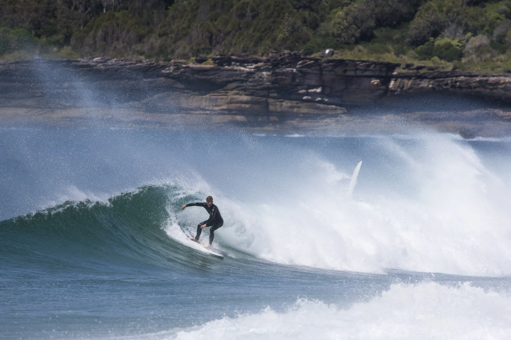 Surfer riding a wave near a rocky shoreline with greenery in the background.