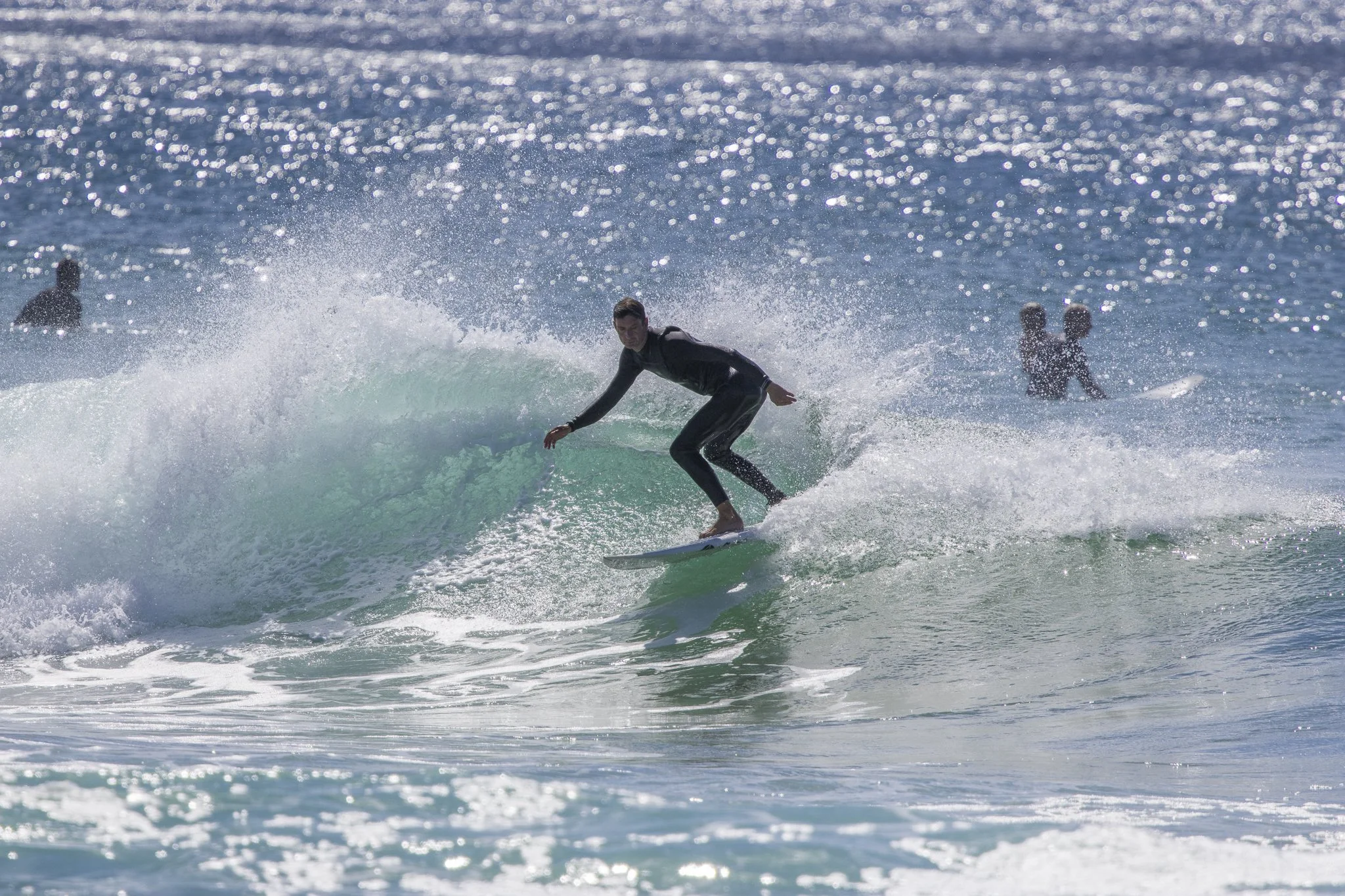 A man surfing on a wave in the ocean with two people in the background, during daytime with sunlight reflecting on the water.