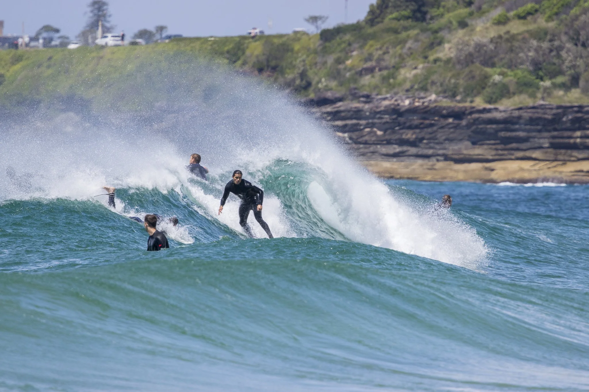 Surfers riding waves on a sunny day at the beach near a rocky coastline, with cars and greenery in the background.
