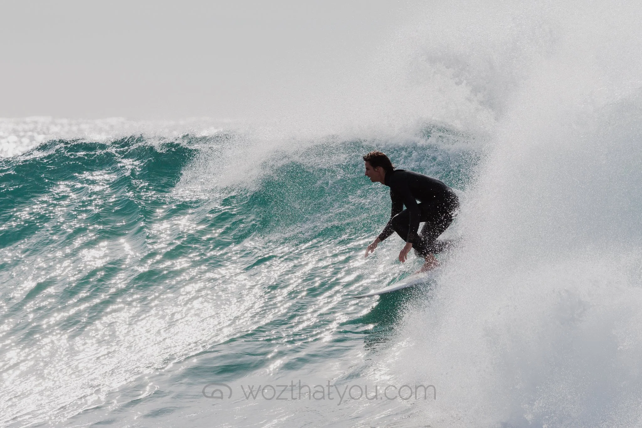 A man surfing on a wave in the ocean, wearing a black wetsuit, crouching on his surfboard, with white spray behind him.