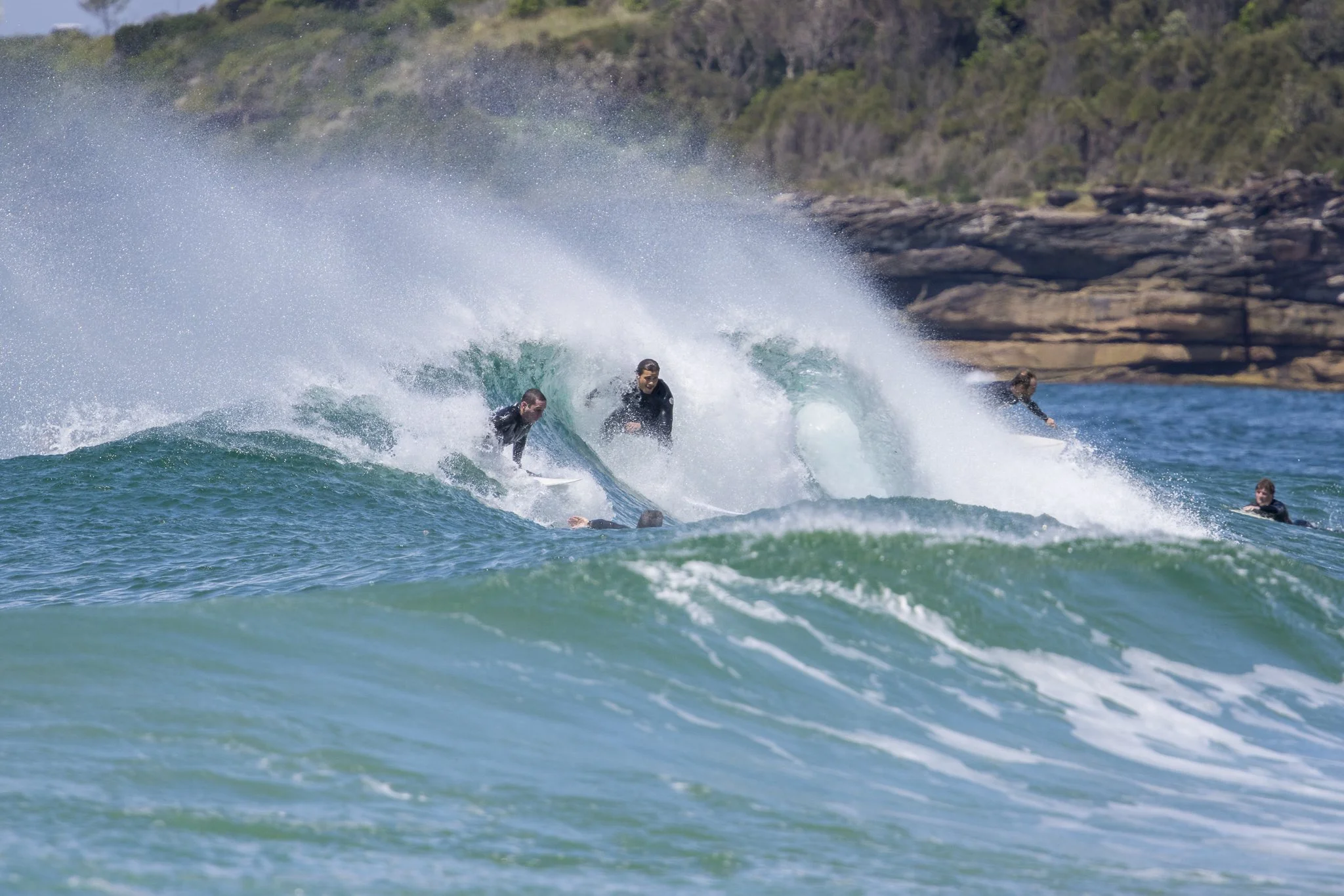 People surfing on a large wave near a rocky coastline with some trees in the background.