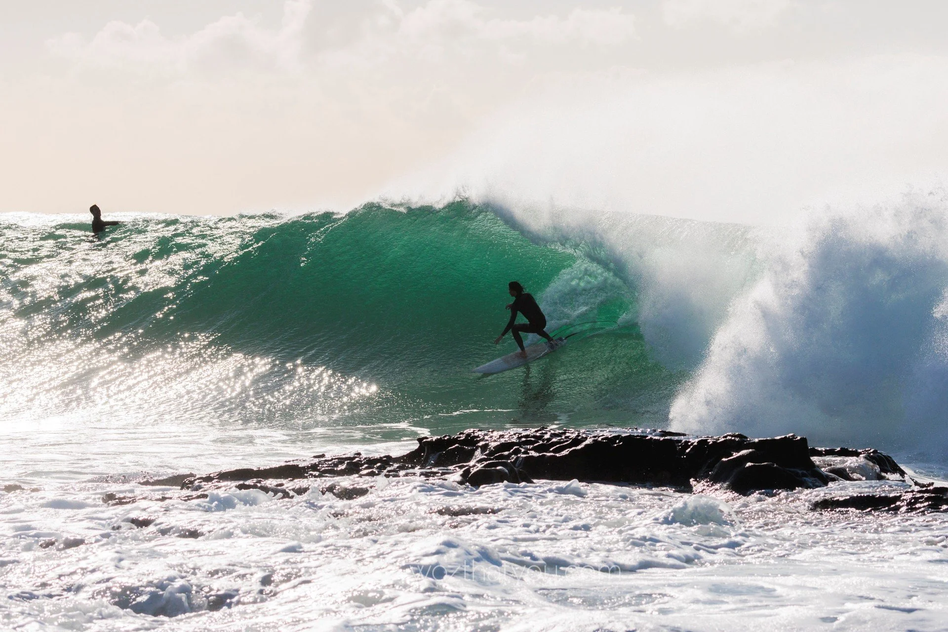 A surfer riding inside the barrel of a large green wave near the shore with foam and rocks in the foreground, and another surfer in the distance.