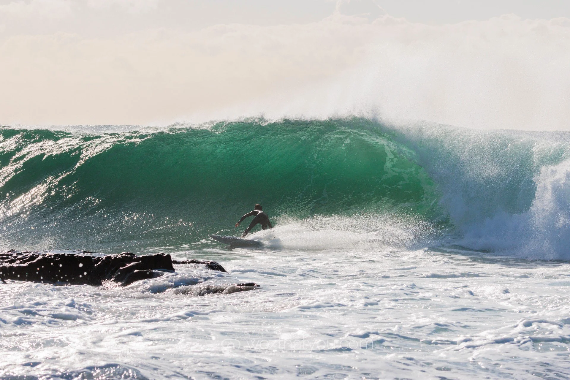 A surfer riding a large ocean wave near rocks on the shoreline
