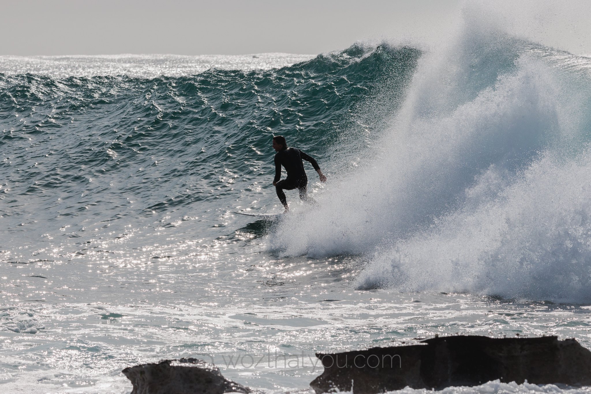 A person surfing on a large ocean wave, wearing a black wetsuit, with a distant horizon and shimmering water in the background.