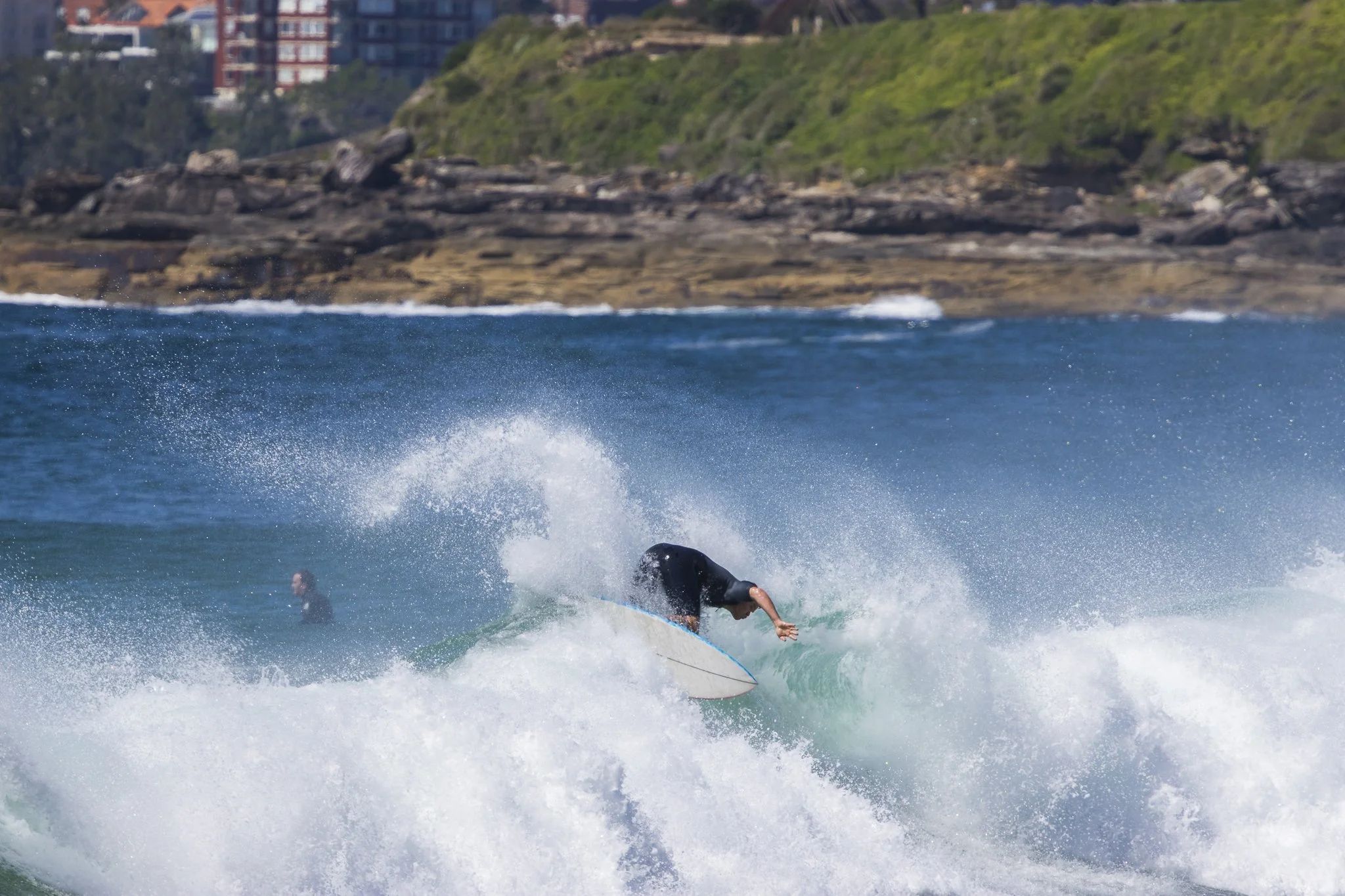 Surfer riding a wave in the ocean near a rocky shoreline with buildings on a hillside in the background.