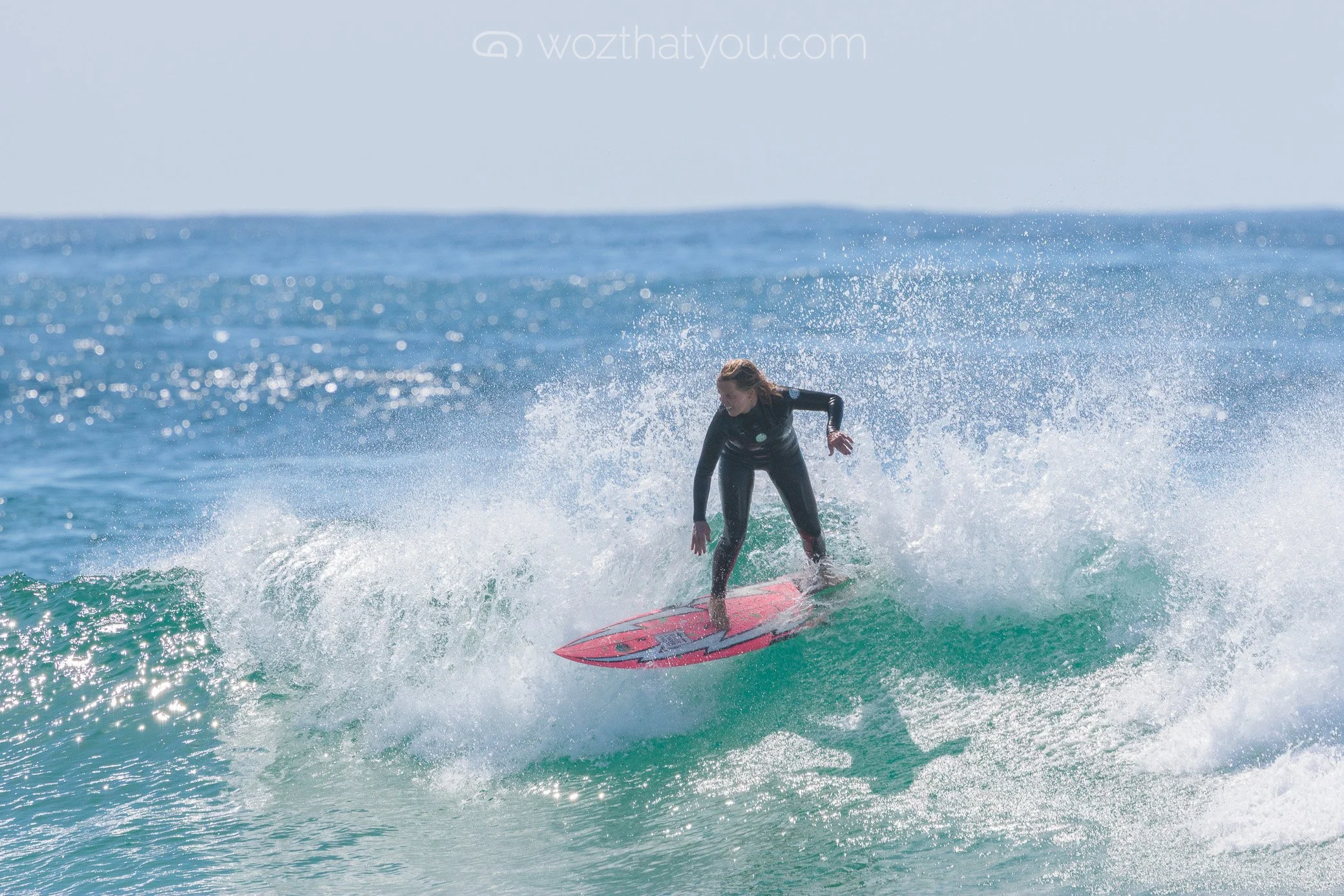A woman surfing on a pink surfboard in the ocean with waves and blue sky.
