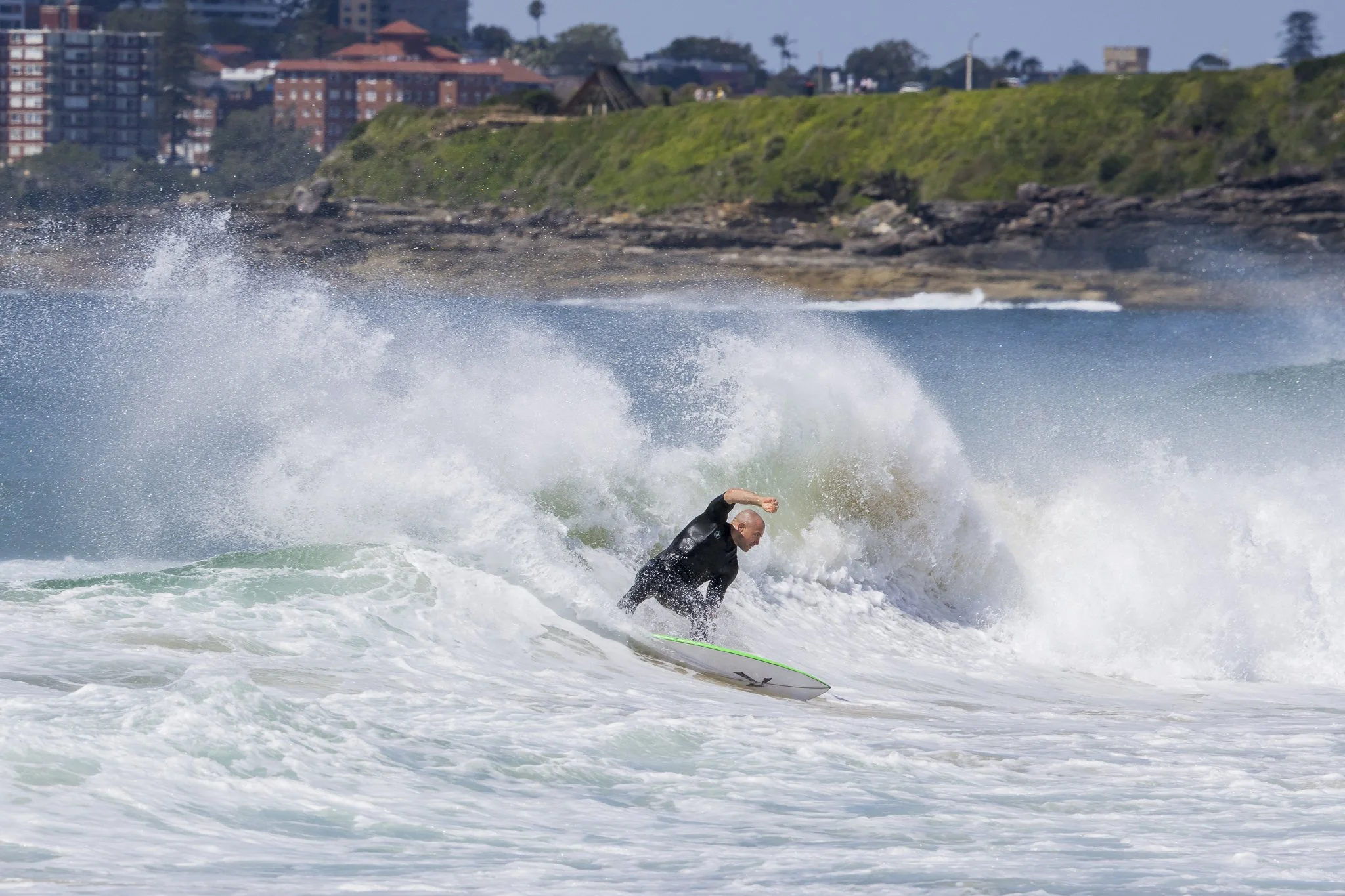 A surfer riding a wave near the shoreline with buildings and green hillside in the background.