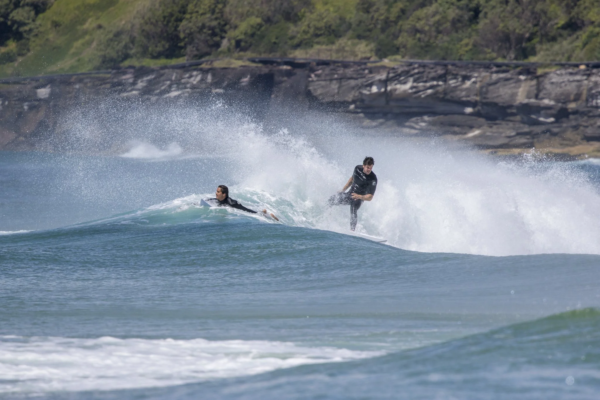 Two surfers riding a wave with a rocky shoreline and green hills in the background.