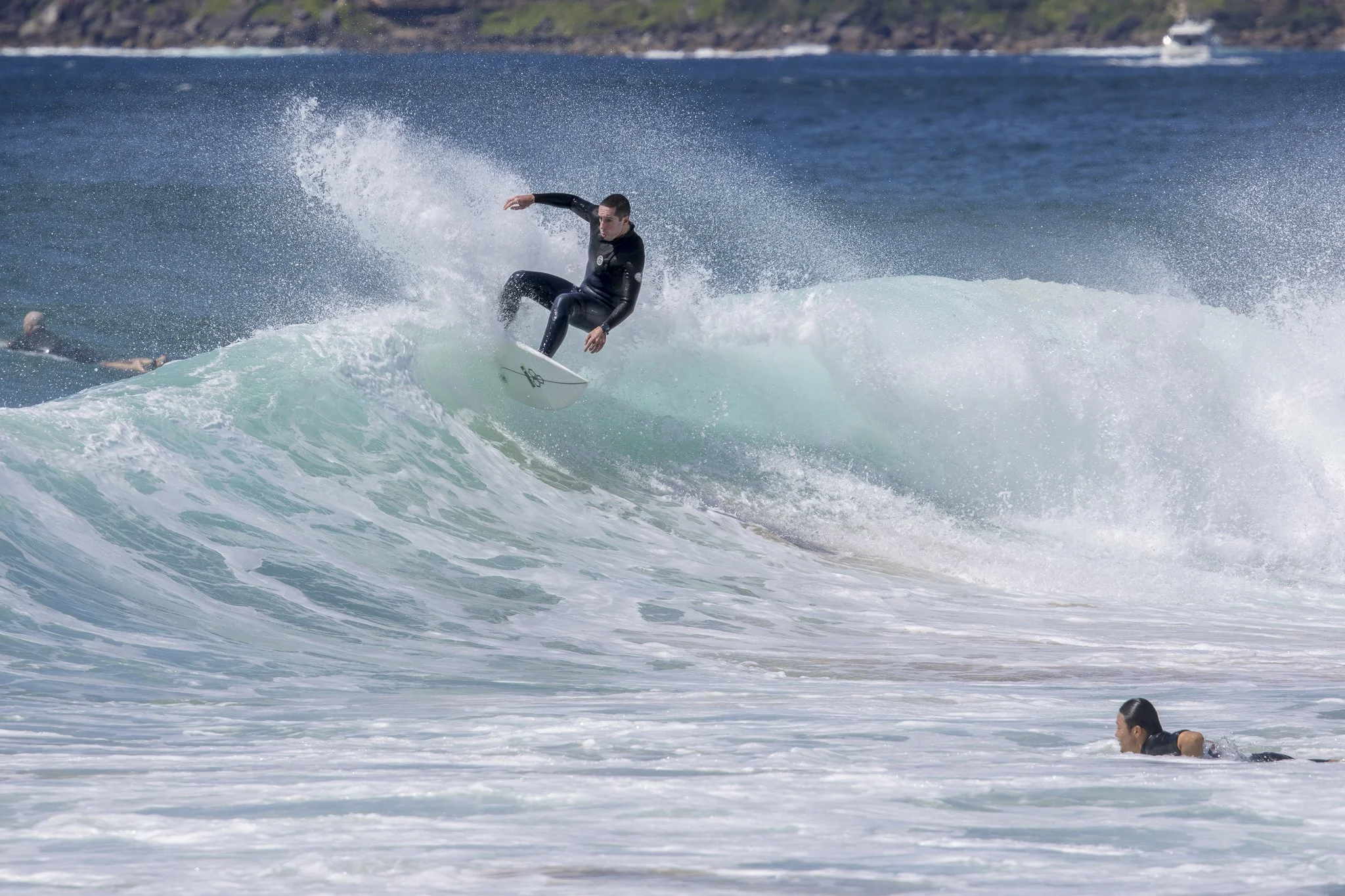 A man surfing on a wave in the ocean, with another person swimming nearby, and a boat in the background.