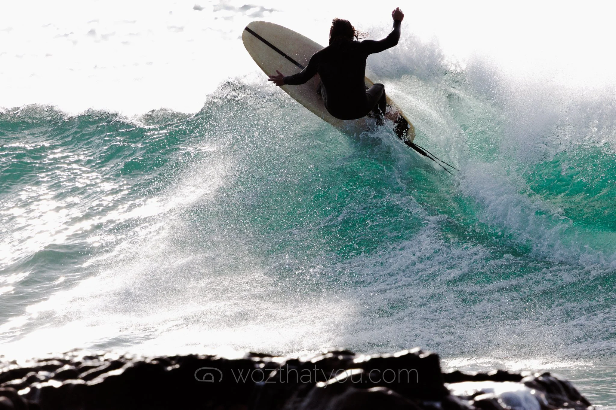 A person riding a wave on a surfboard, mid-air, wearing a wetsuit, with ocean spray around them.