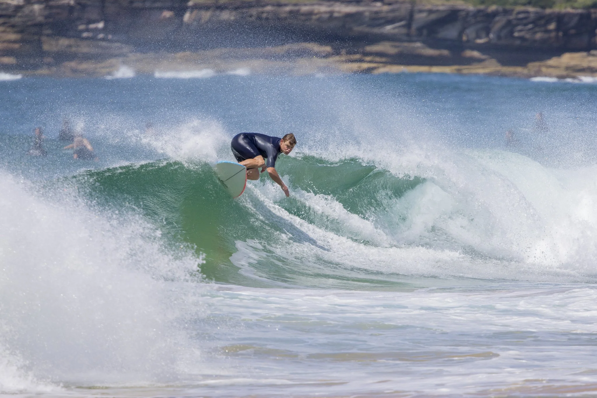 A male surfer in a black wetsuit riding a green wave at the beach, with other people in the water in the background and rocky coastline in the distance.