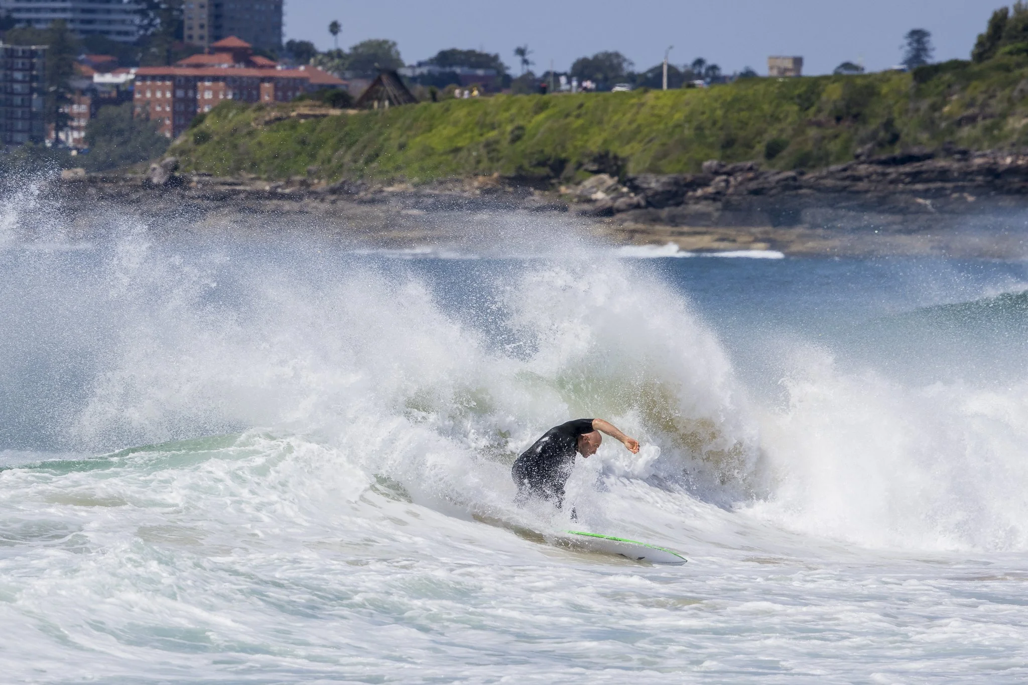 A person surfing on a wave near a rocky shoreline with buildings and trees in the background.