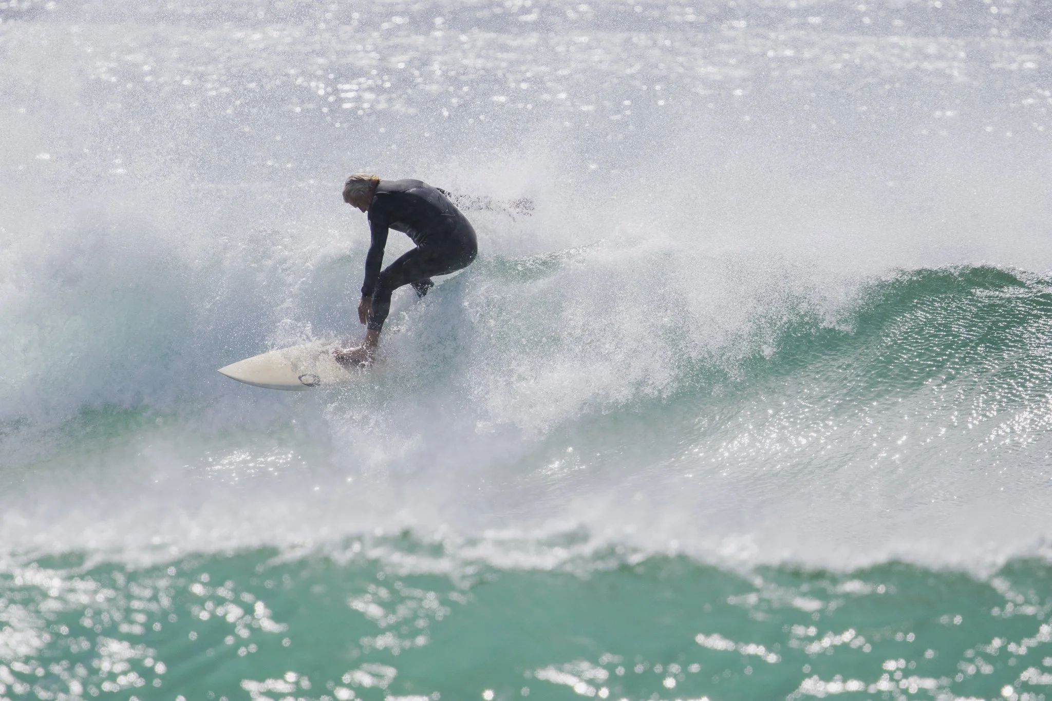 A person surfing on a wave, dressed in a black wetsuit, riding a white surfboard in the ocean.