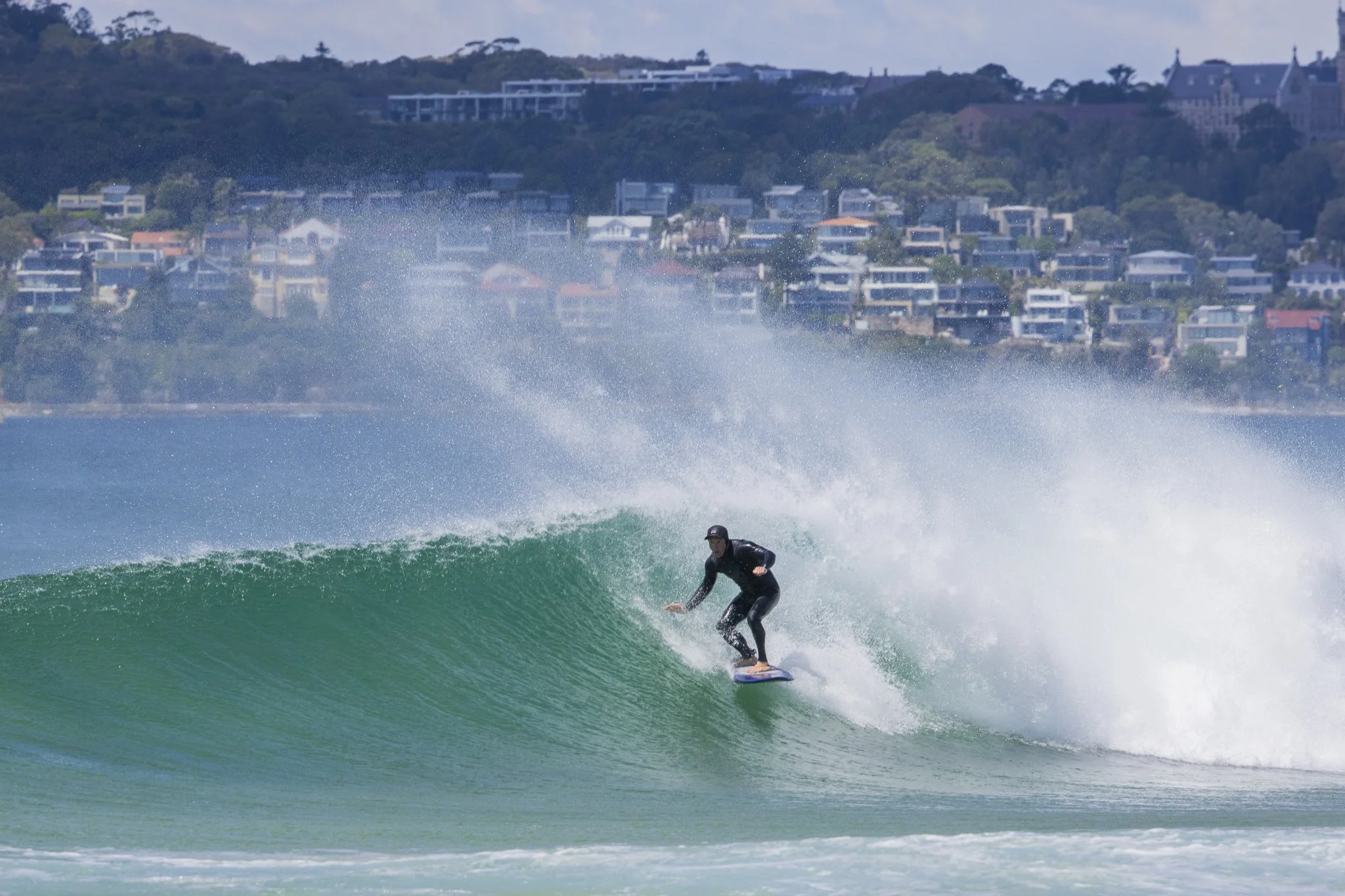 Person surfing on a wave near a coastal town with houses on a hillside in the background.