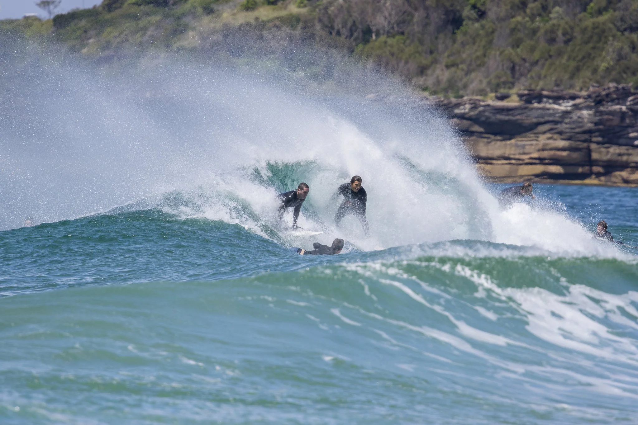 Surfers riding a wave at the beach with a rocky shoreline and green hillside in the background.
