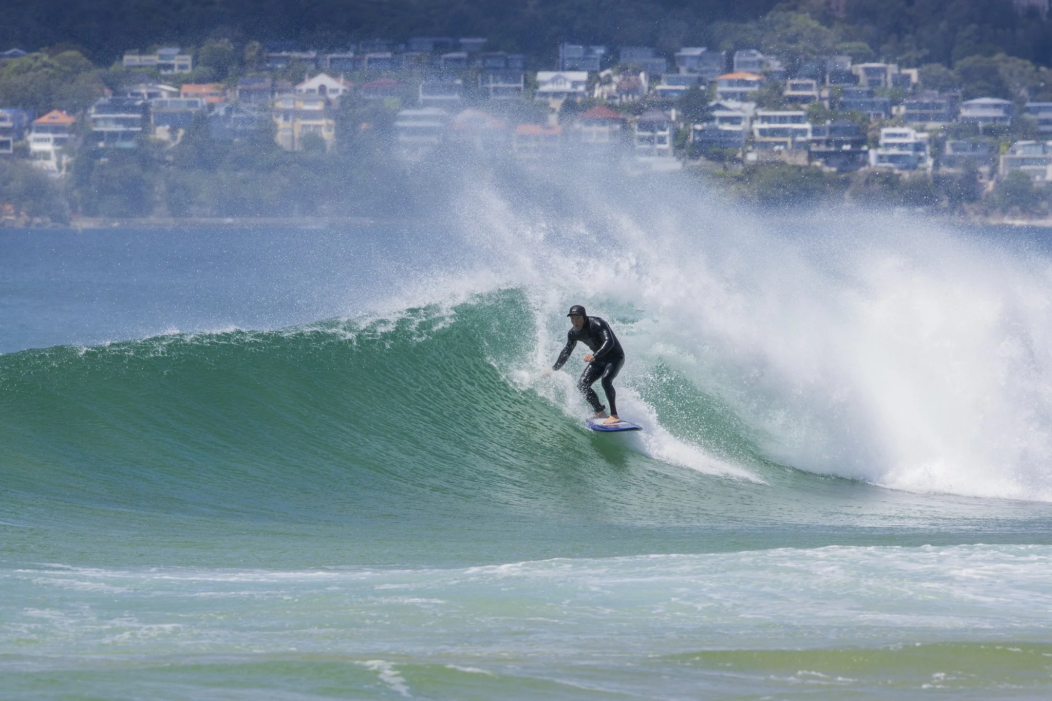 A person in a black wetsuit is surfing on a wave with a cityscape and hillside houses in the background.