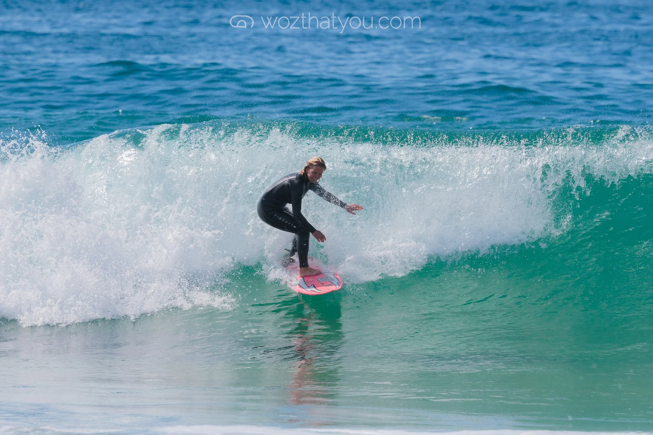 A woman surfing on a pink surfboard in blue ocean waves, wearing a black wetsuit.