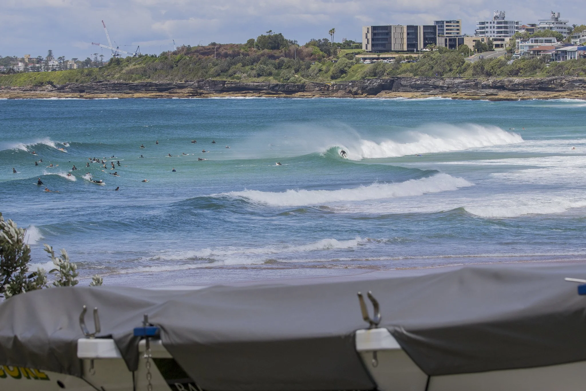 Surf beach with many surfers in the water, waves breaking, and a cityscape on the hillside in the background.