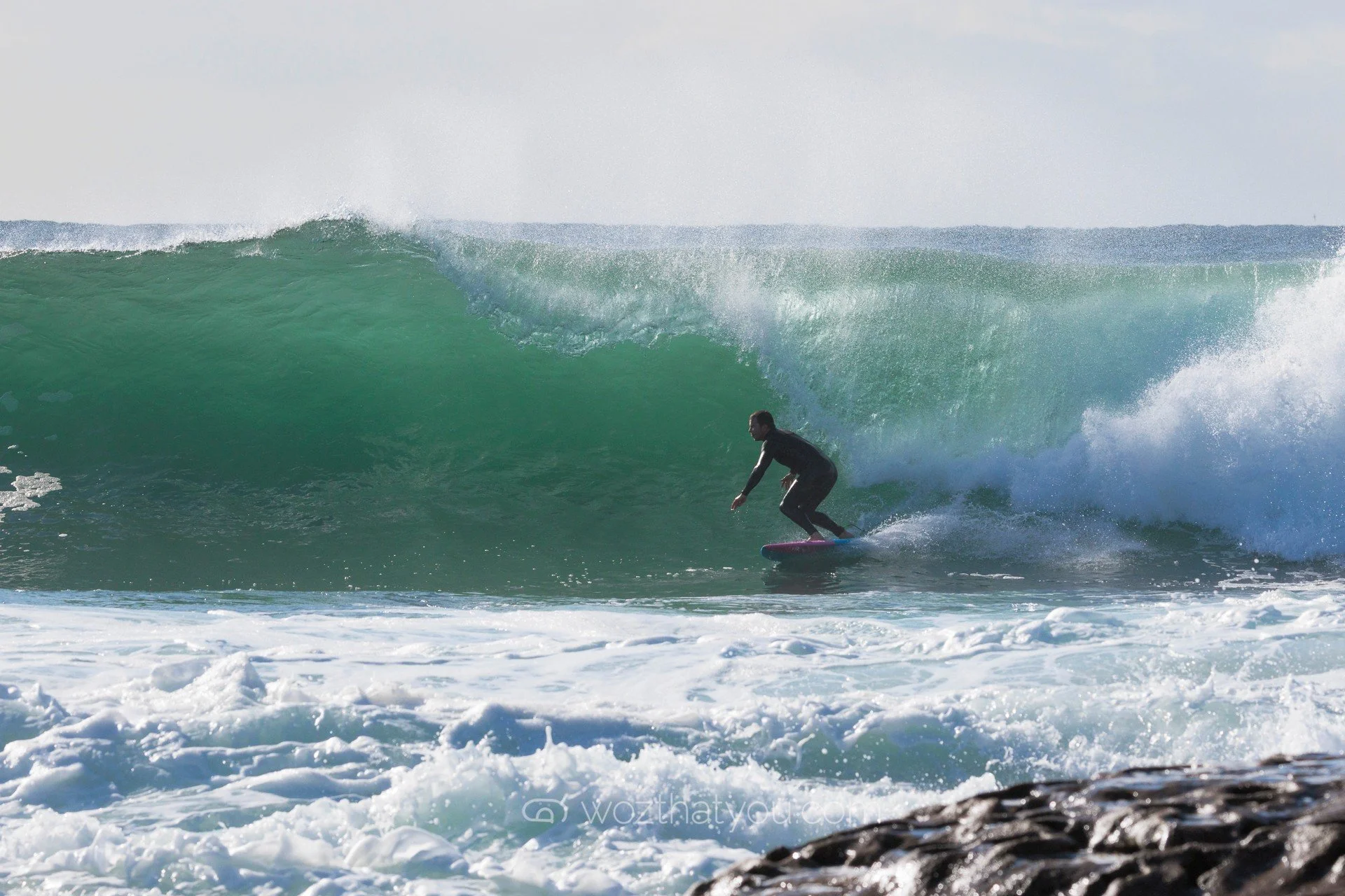 Person surfing on a large green wave with rocky shoreline in the foreground.