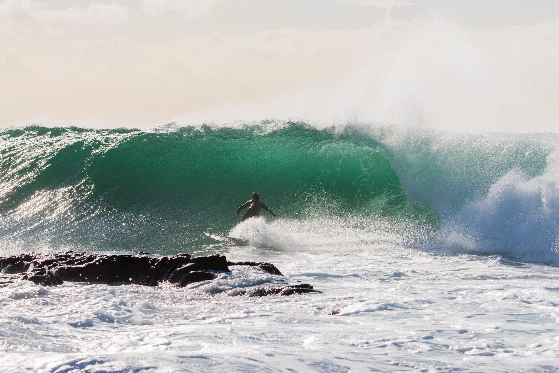 A person surfing on a large green wave near rocky shoreline, with foam in the foreground.