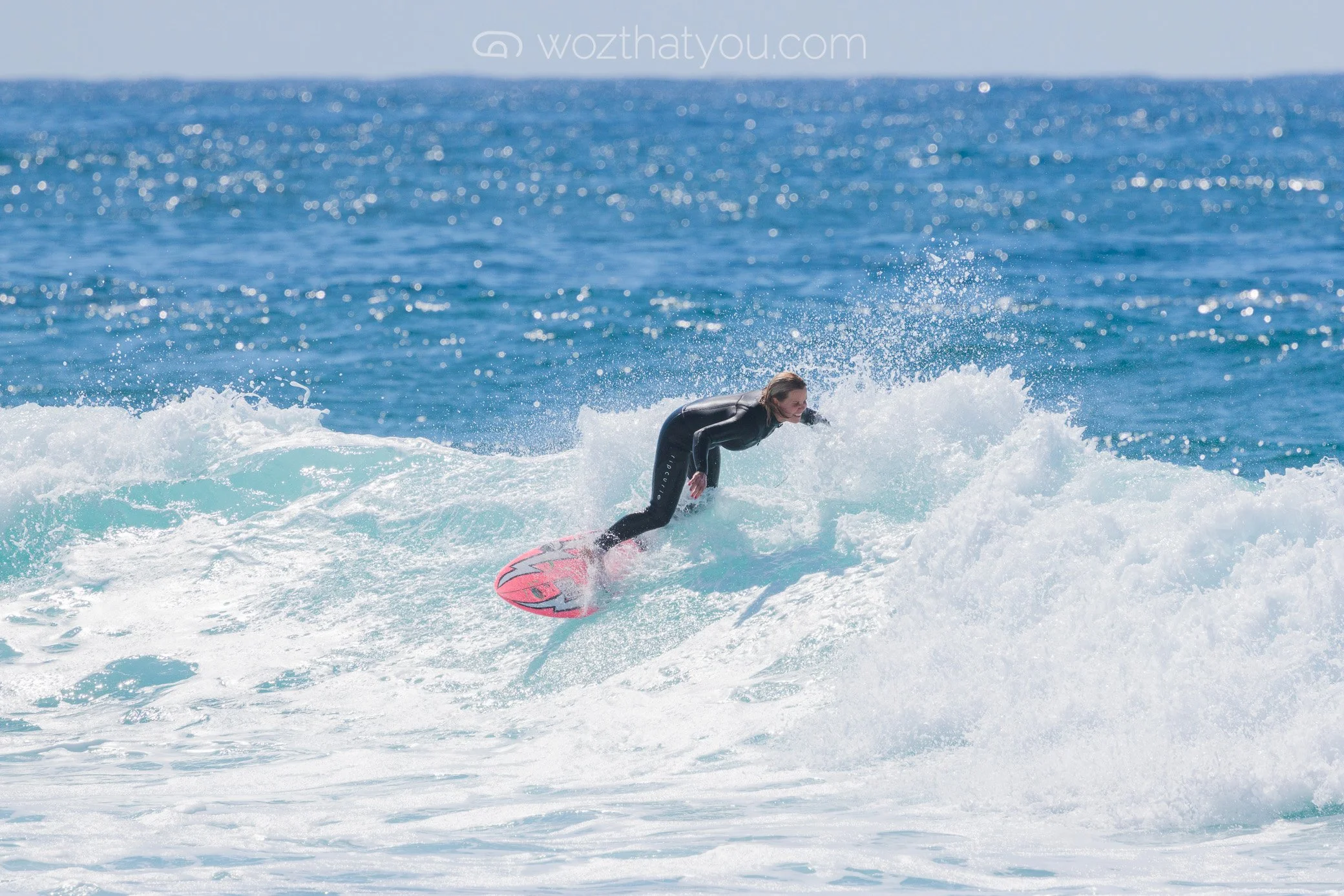 A woman surfing on a pink surfboard in the ocean with blue water and white waves.