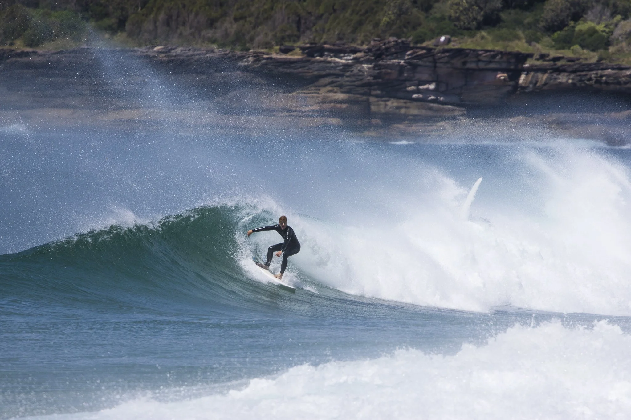 A person surfing on a wave in the ocean with a rocky shoreline and green trees in the background.