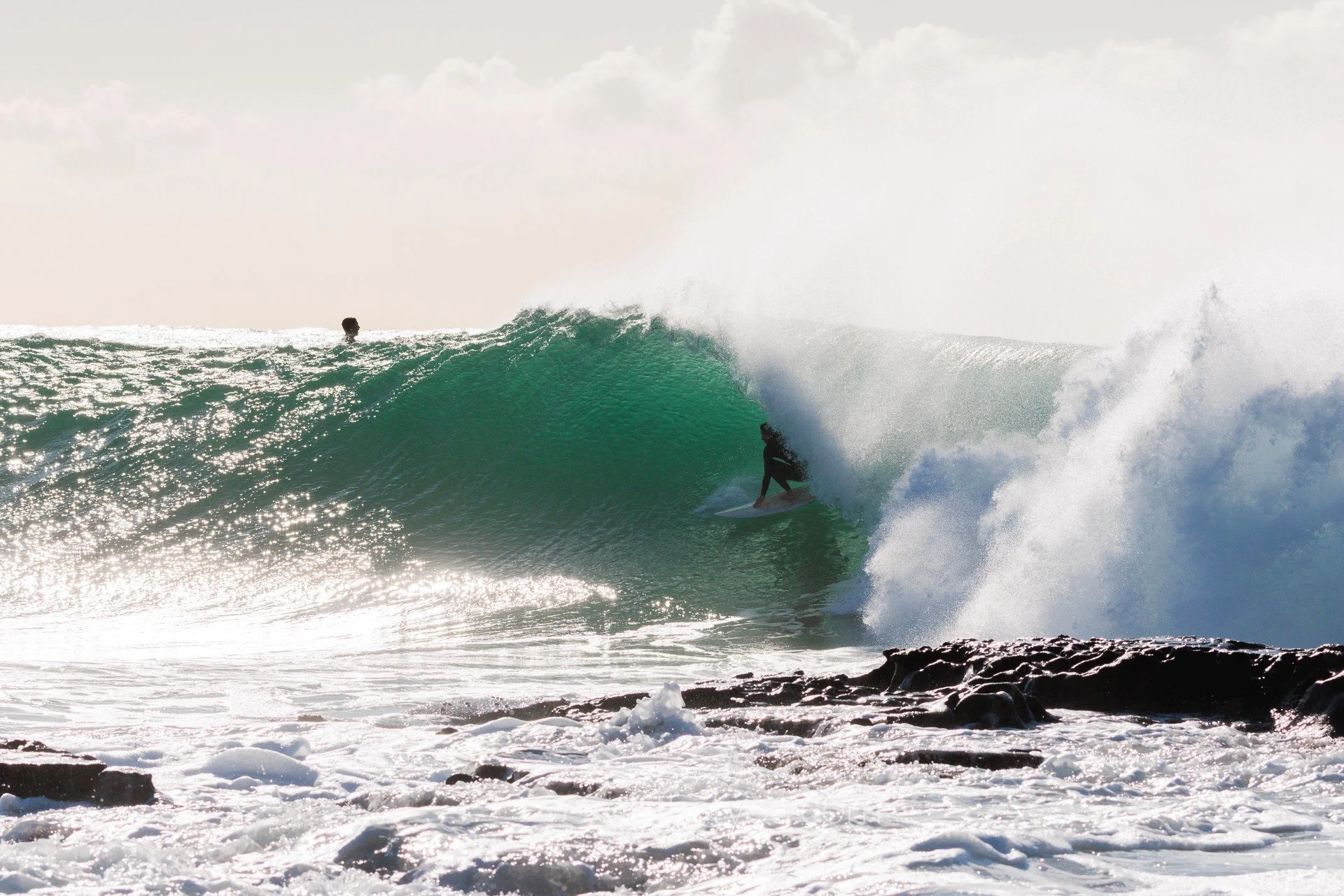 A person surfing on a large wave near rocks, with another person watching in the background.