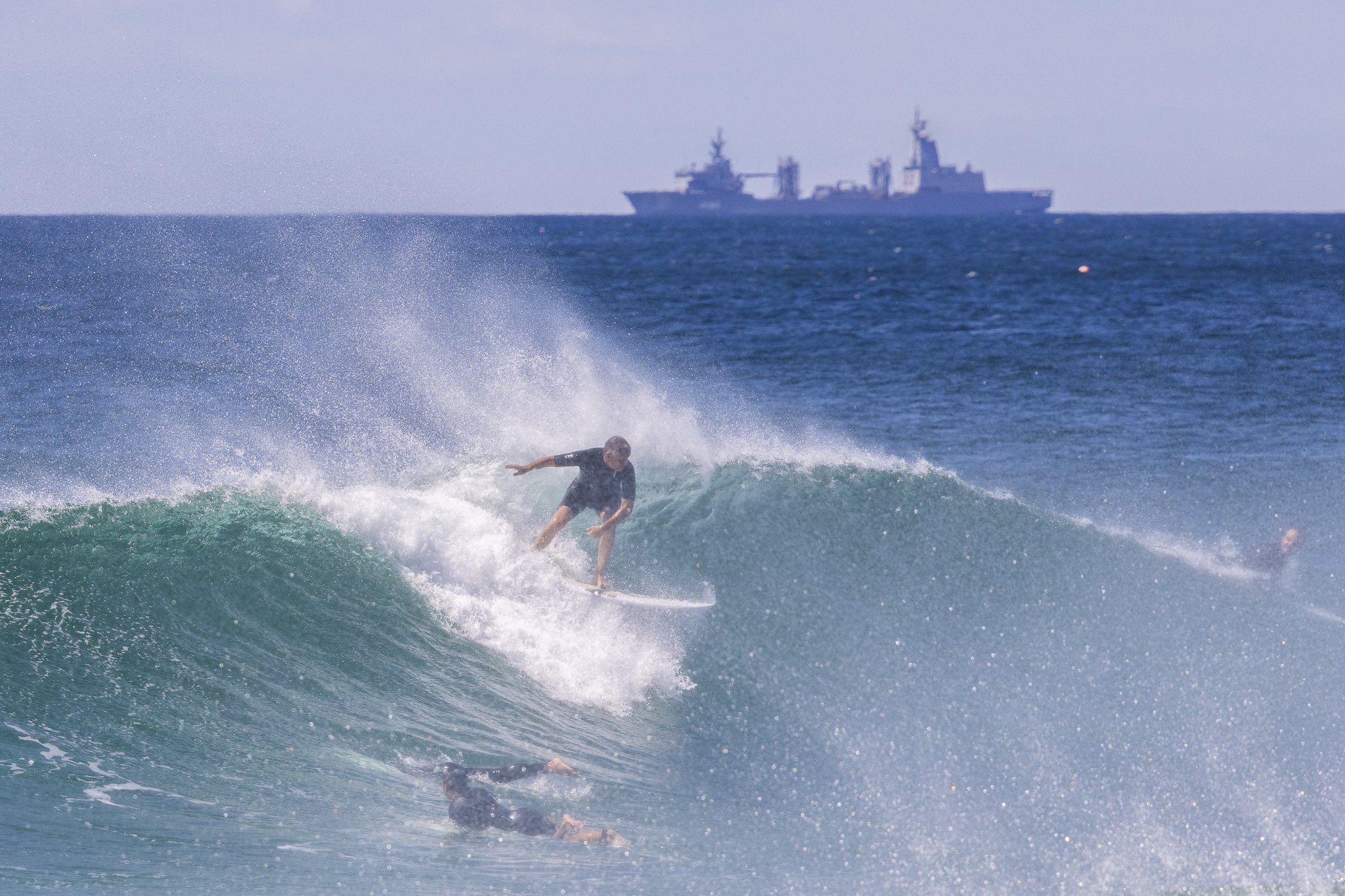 A person surfing on a wave with another surfer in the water below. A large ship is visible in the distance on the ocean horizon.