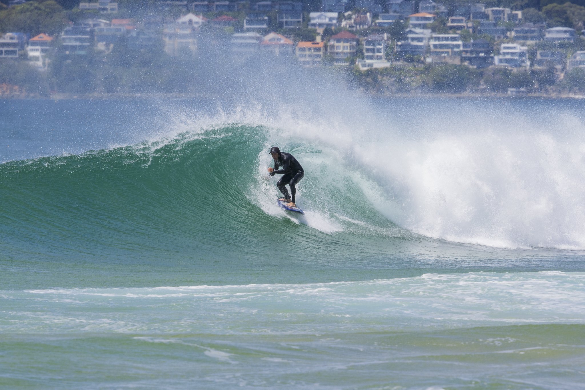 A person surfing on a wave in the ocean with a backdrop of houses on a hillside.