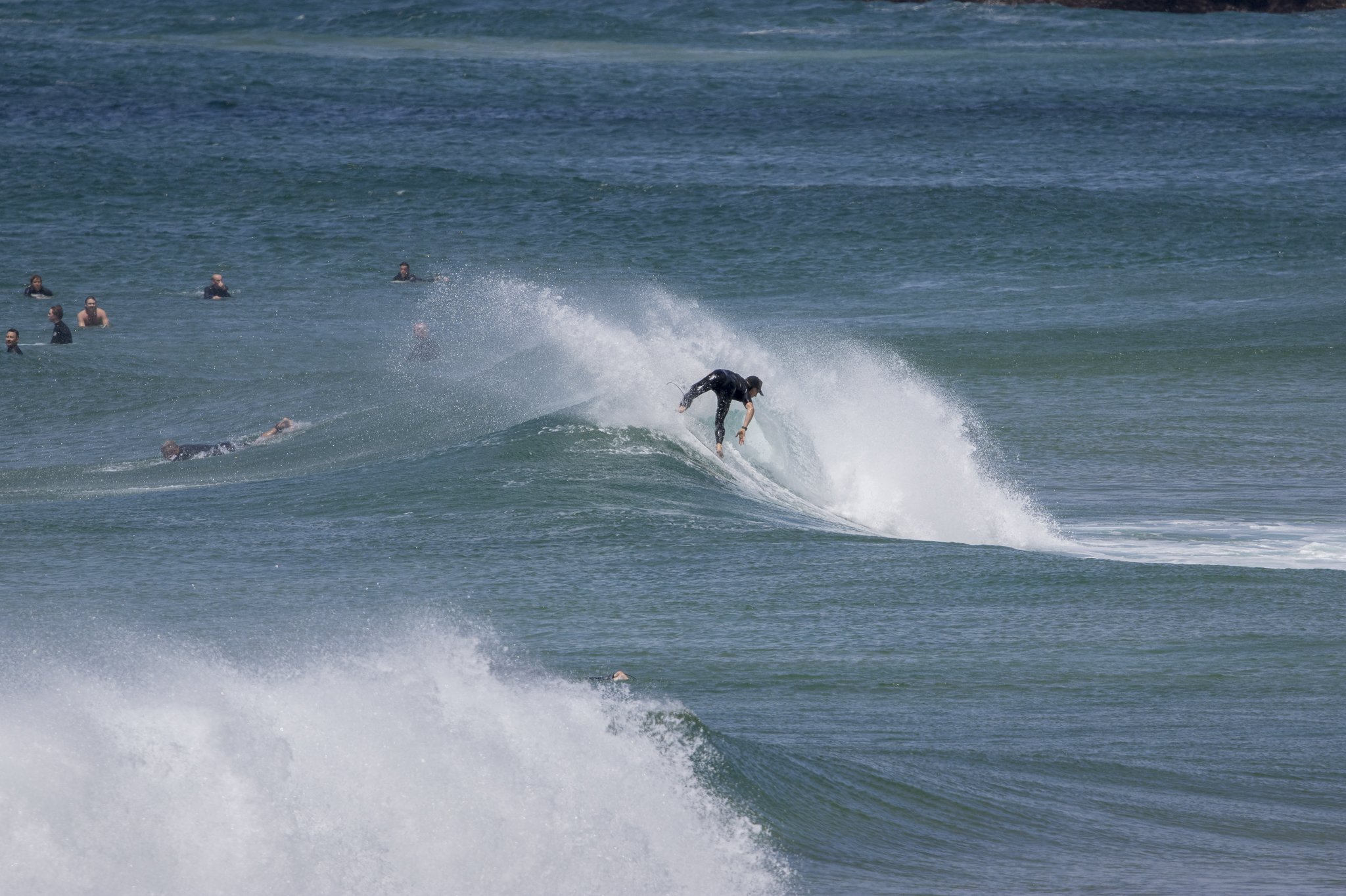 A person in black wetsuit surfing on a wave in the ocean, with several other people swimming and floating in the water in the background.