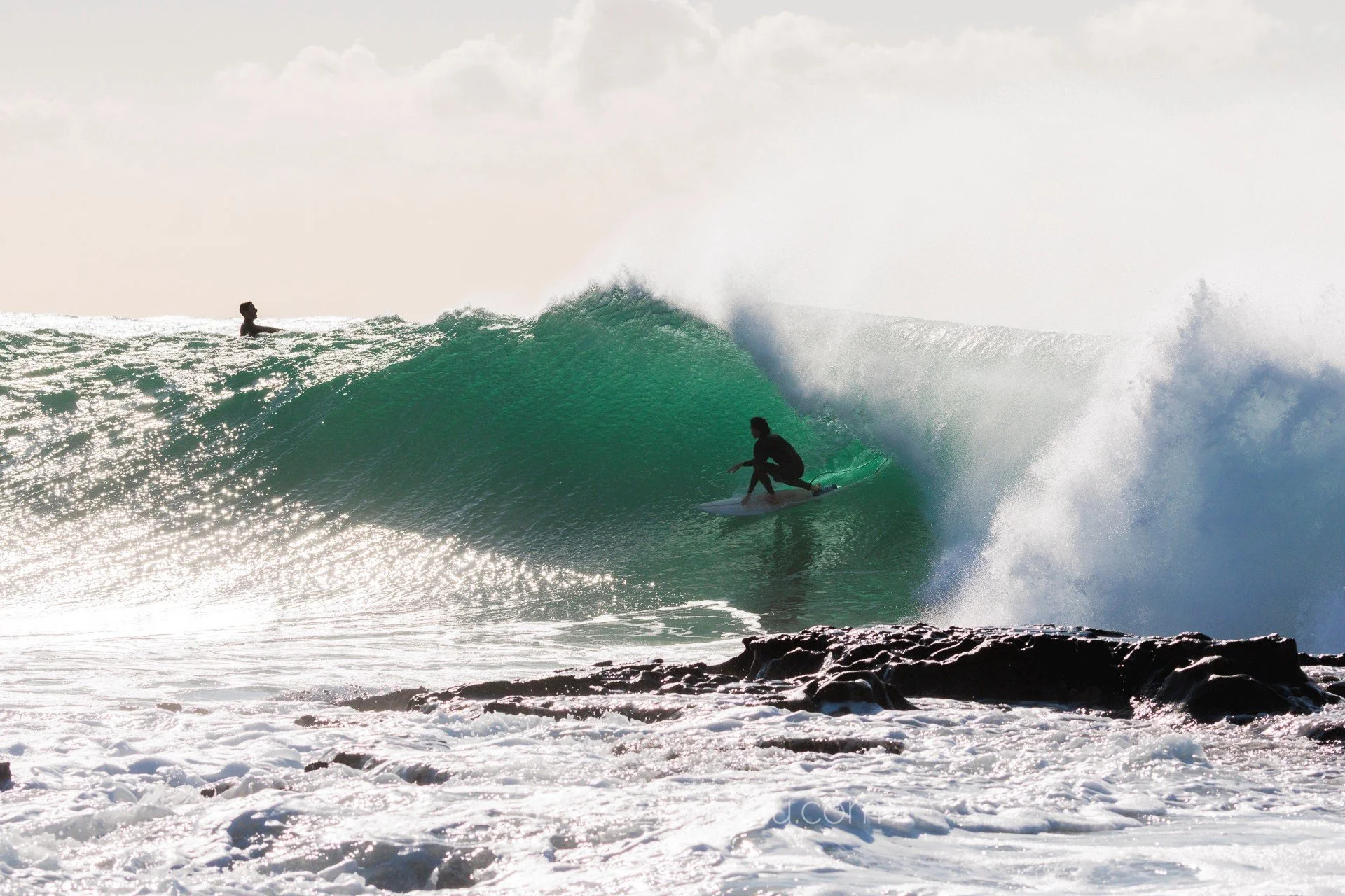 A surfer in a black wetsuit riding inside the barrel of a large green wave, with another surfer watching from the top of the wave and a rocky shoreline in the foreground.