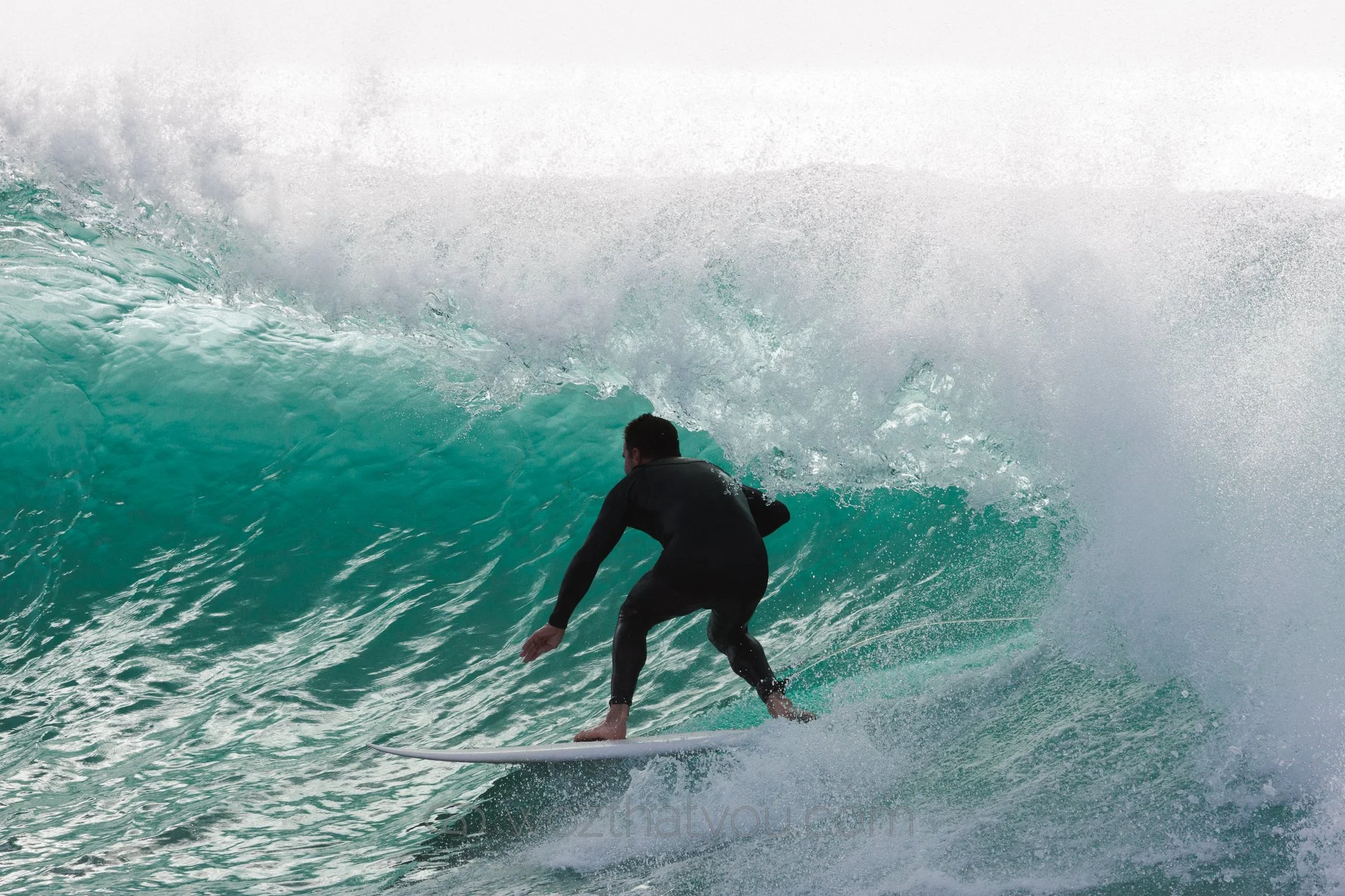 A male surfer in a black wetsuit riding a large turquoise wave.