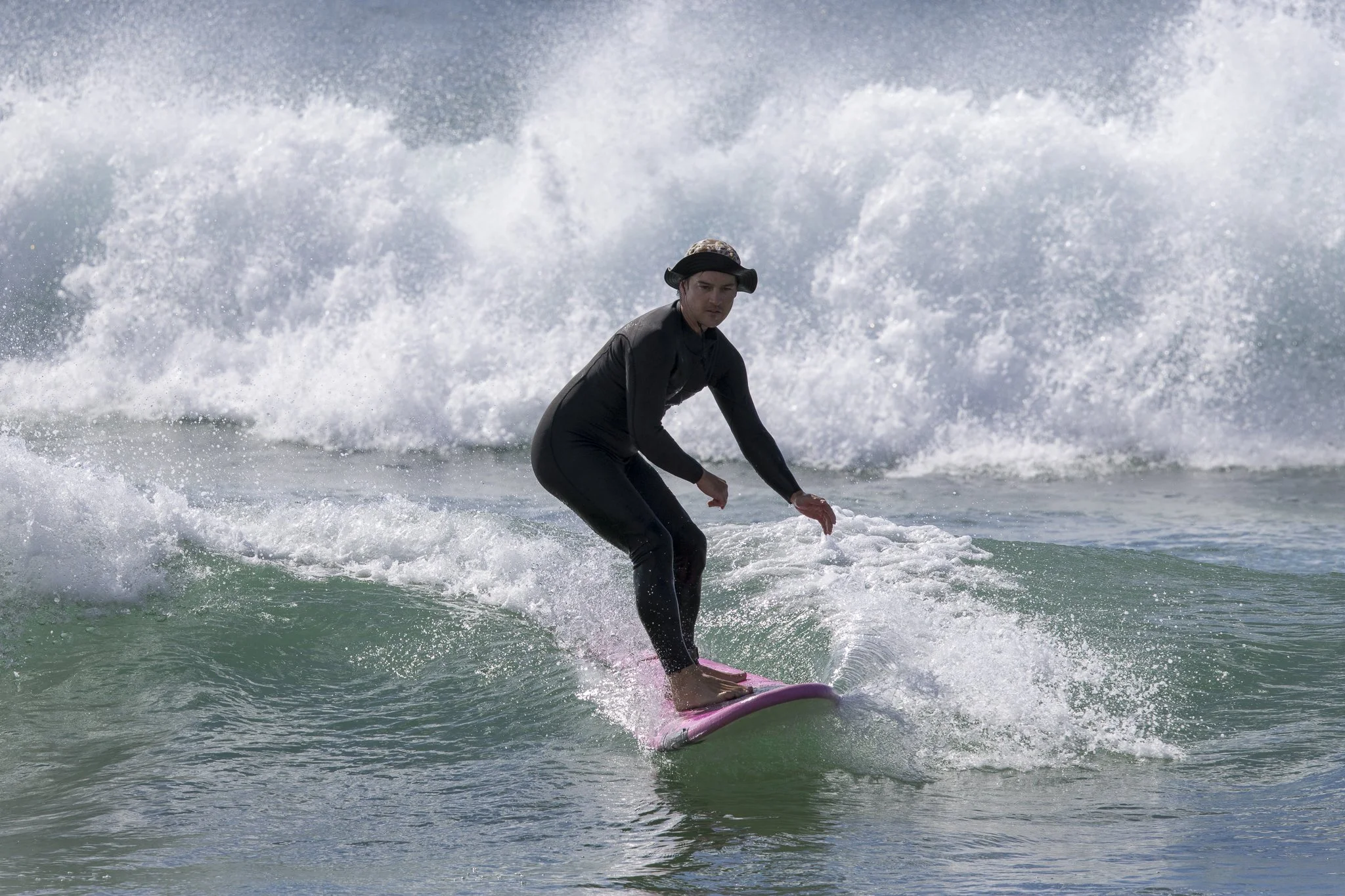 A person wearing a black outfit and hat surfing on a pink surfboard on the ocean.