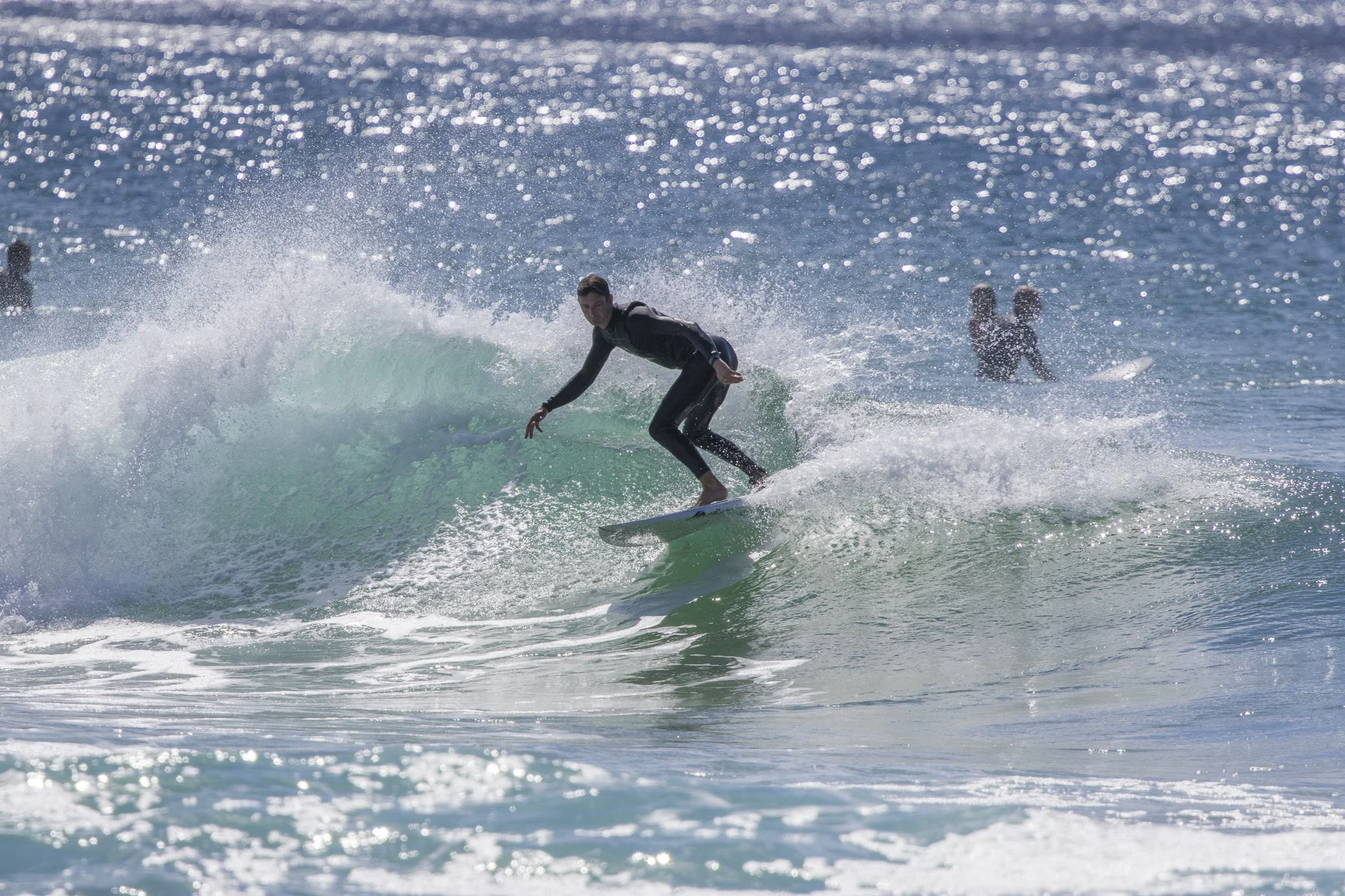 A surfer riding a wave in the ocean with other surfers in the background, sunlight sparkling on the water.