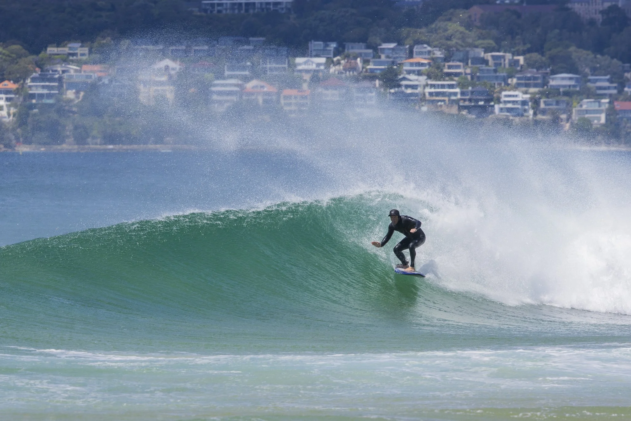 A person surfing on a wave in the ocean with houses on a hillside in the background.