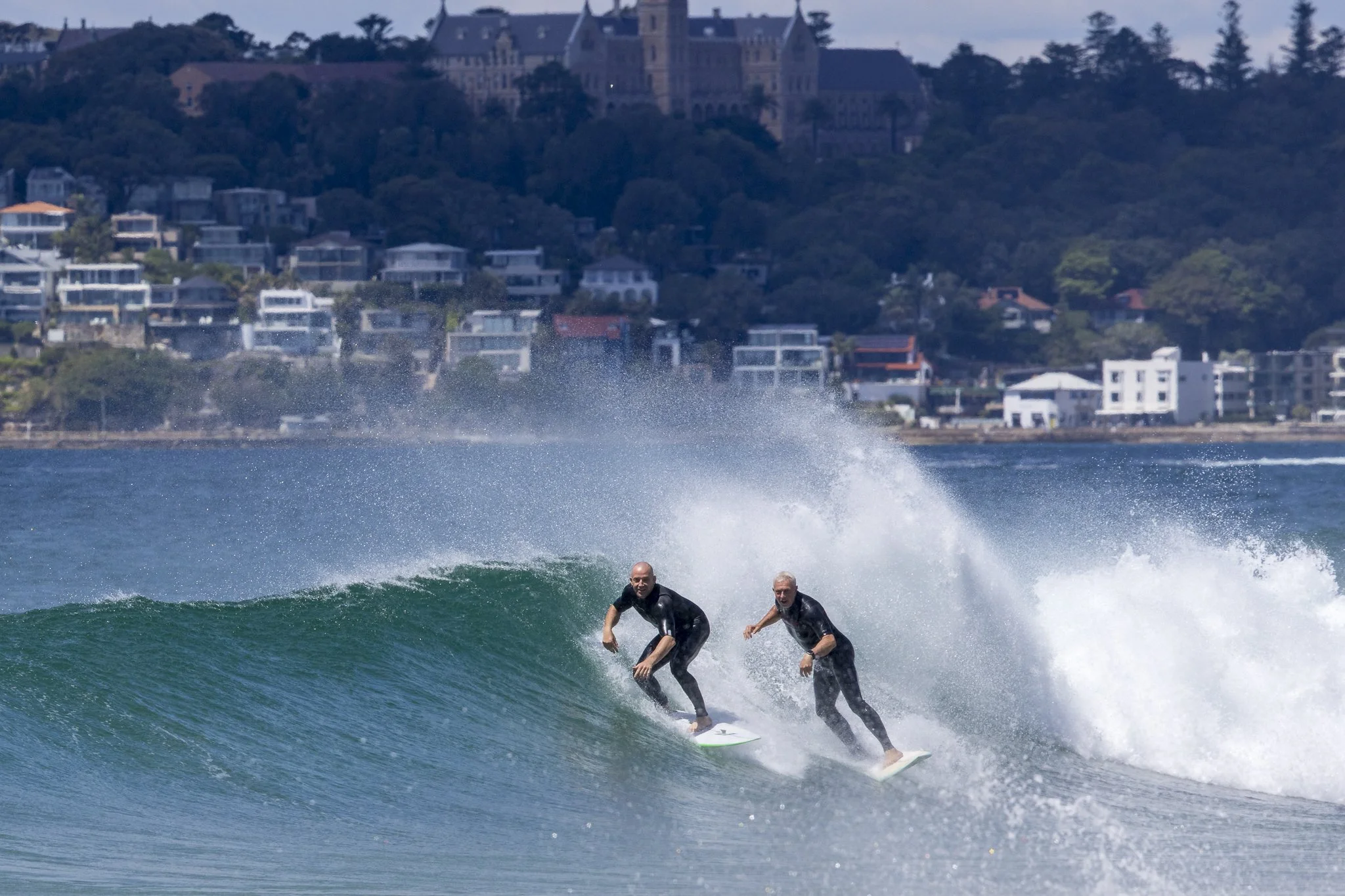 Two surfers riding a wave near the shore, with a cityscape and a large building on a hill in the background.