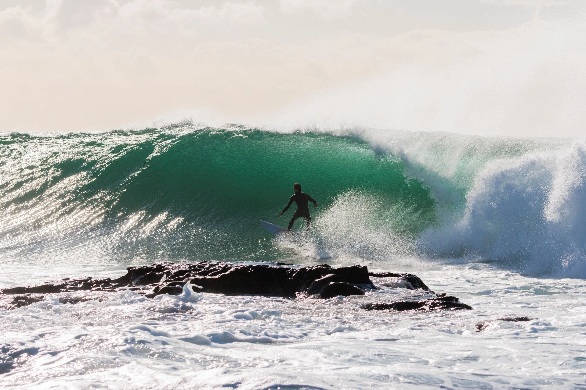 A person surfing on a large green wave near rocks with white foam at the shoreline.